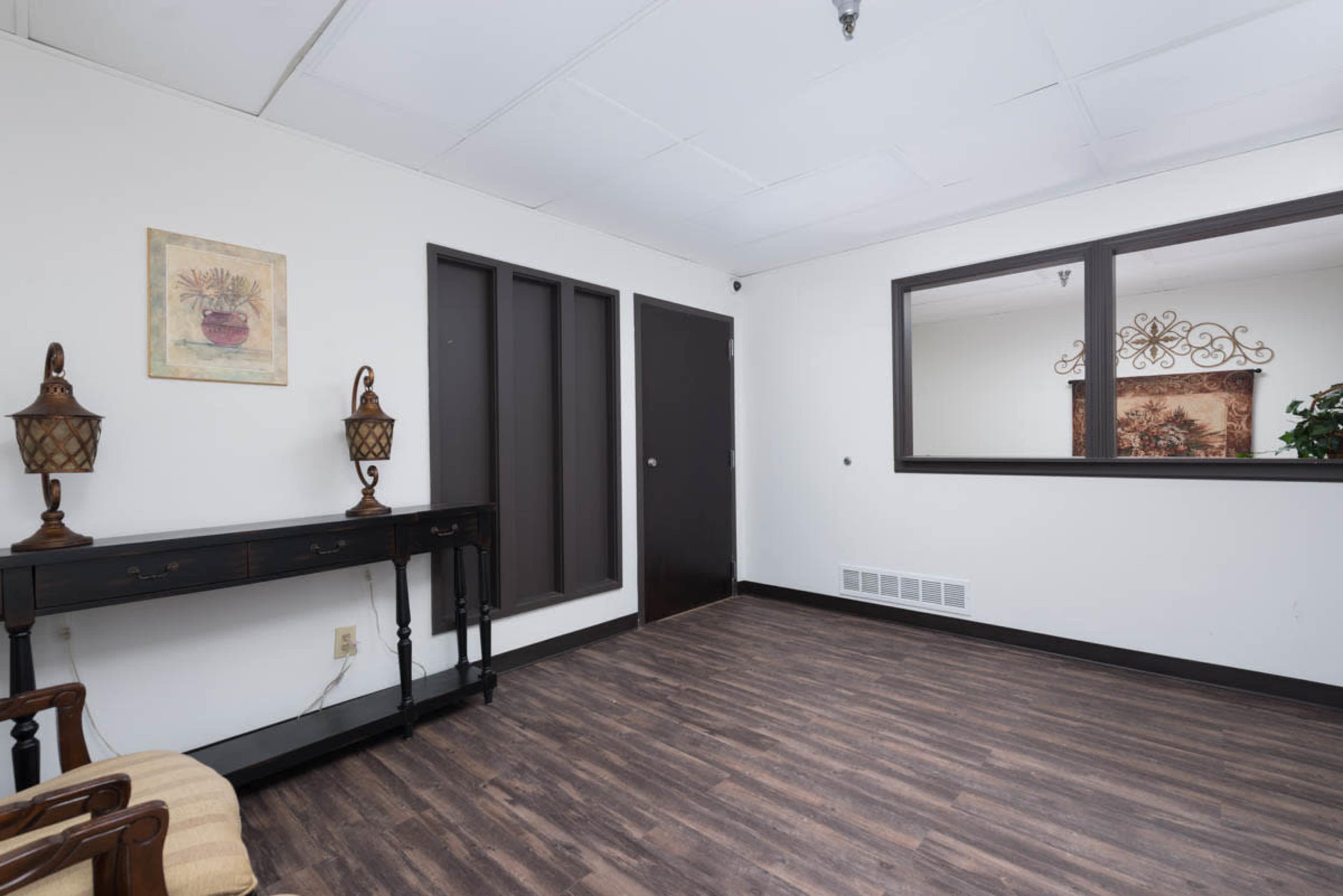 The image shows a sparsely furnished interior room featuring a dark wooden console table, a chair, and framed wall art.