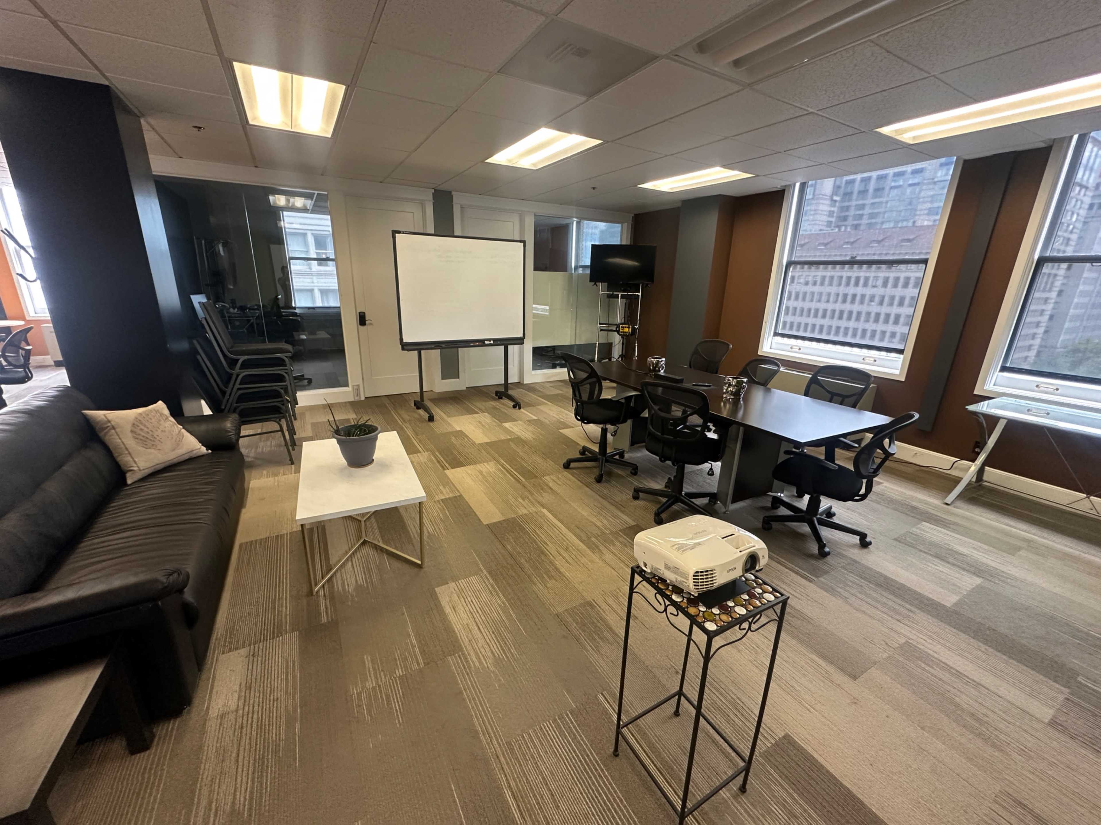 The image shows a modern office meeting space featuring a conference table, chairs, a whiteboard, and a couch, with large windows allowing natural light.