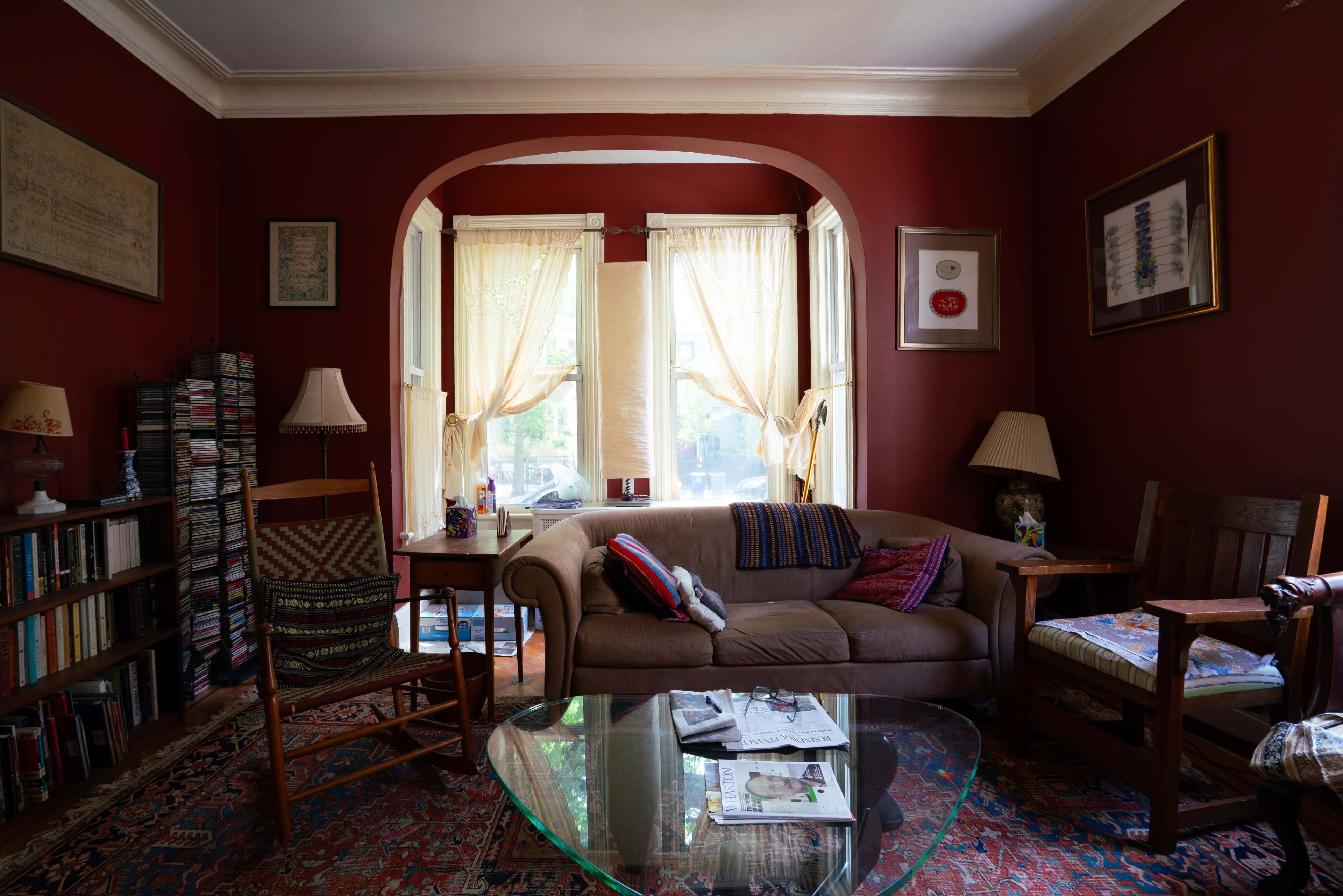 The image depicts a cozy living room with red walls, a brown sofa, a glass coffee table, and various bookshelves and artwork.