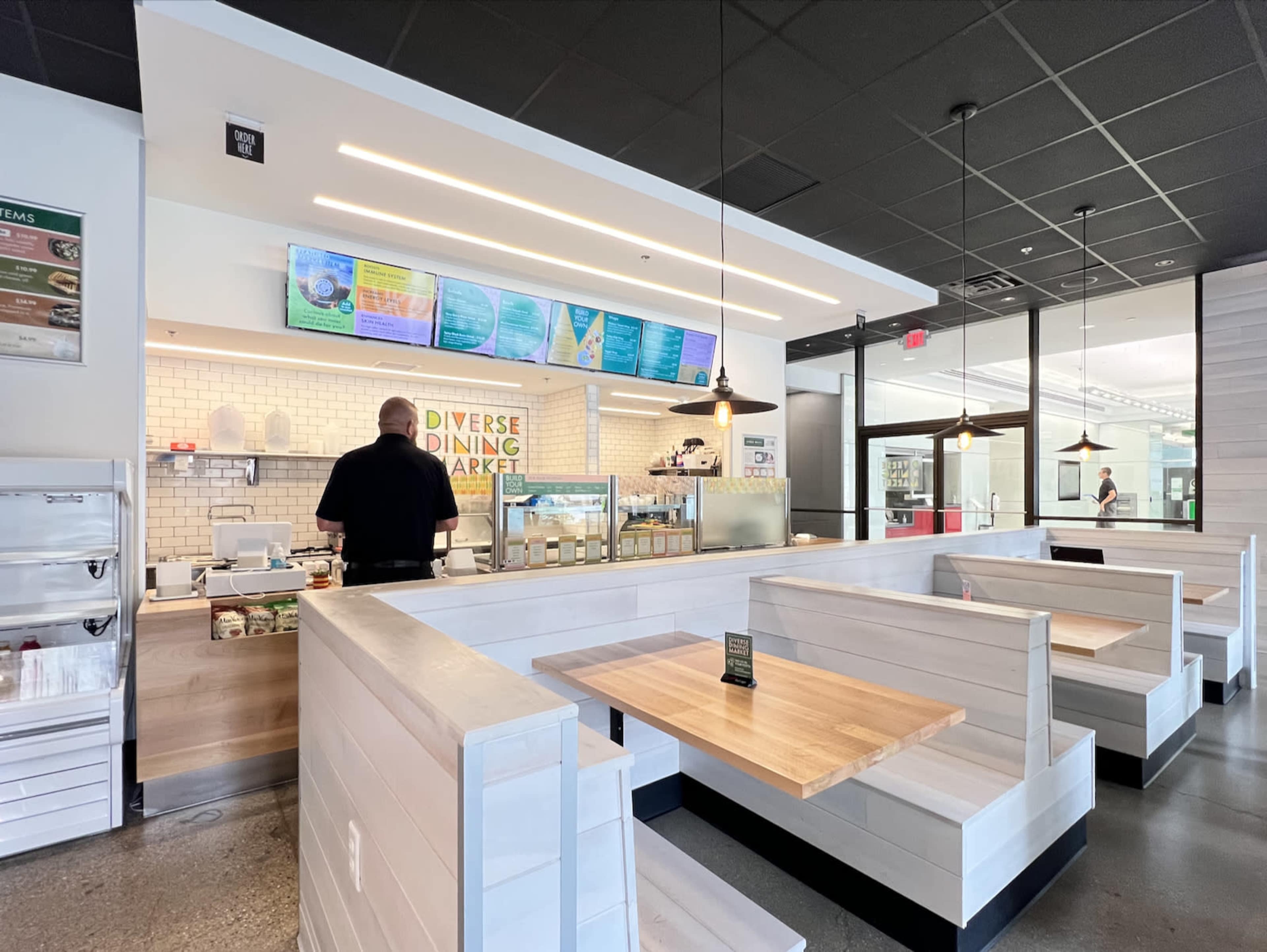The image shows a modern dining area with white and wooden seating, featuring a counter and menu displays in a well-lit restaurant.