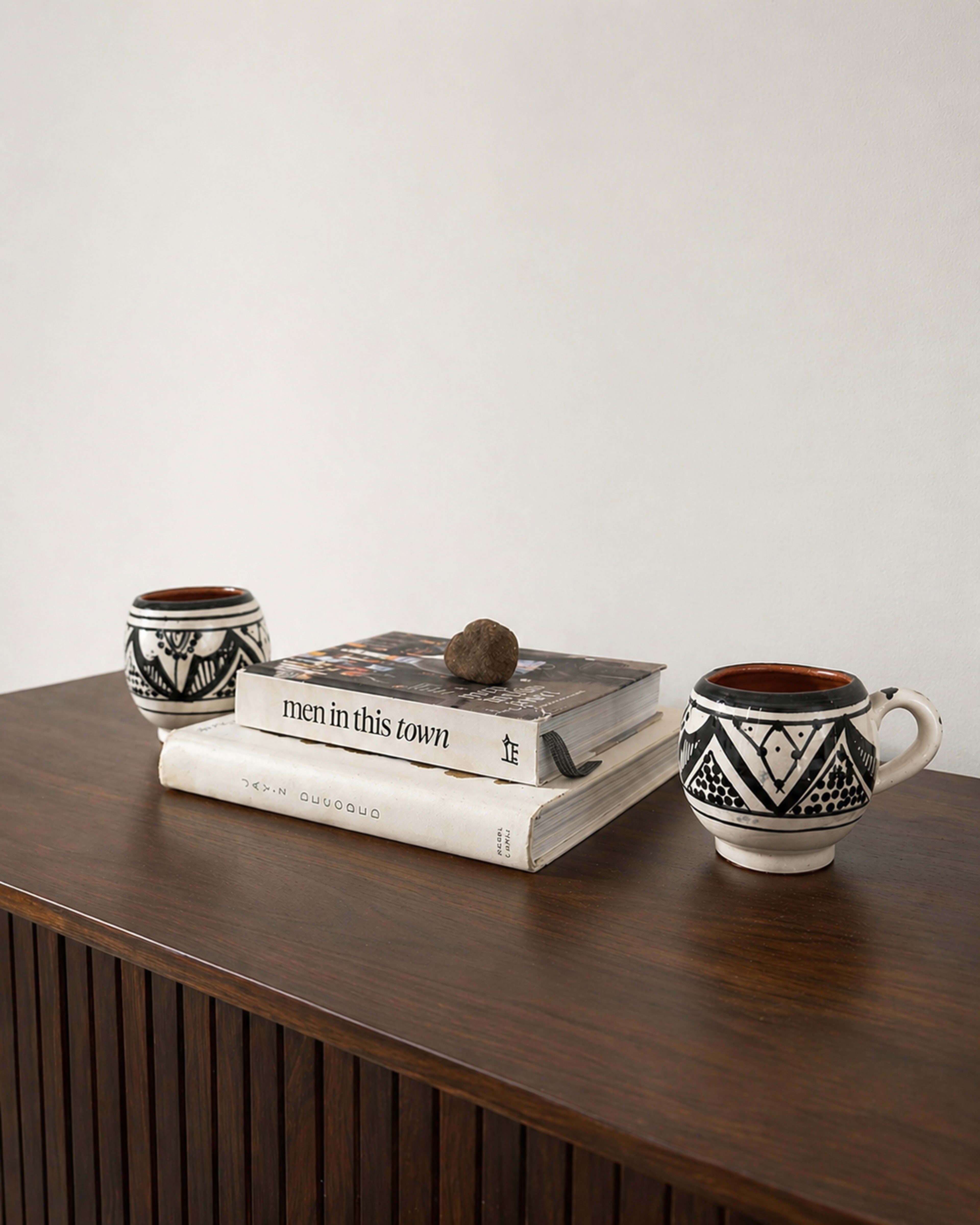 The image shows a wooden table with two ceramic mugs, stacked books, and a decorative stone on top.