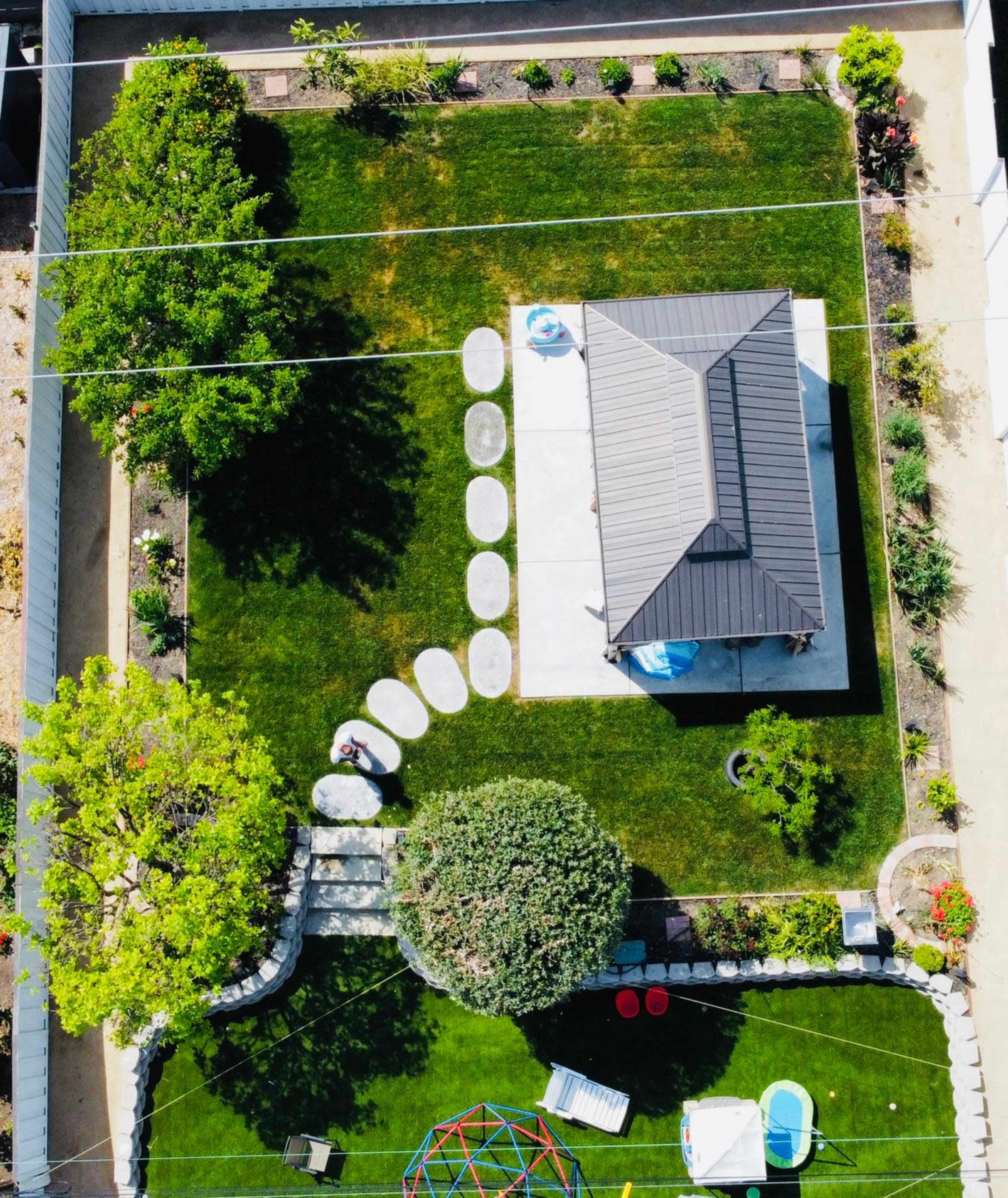 The image shows an aerial view of a landscaped backyard featuring a small house, stone pathway, and various plants and play equipment.