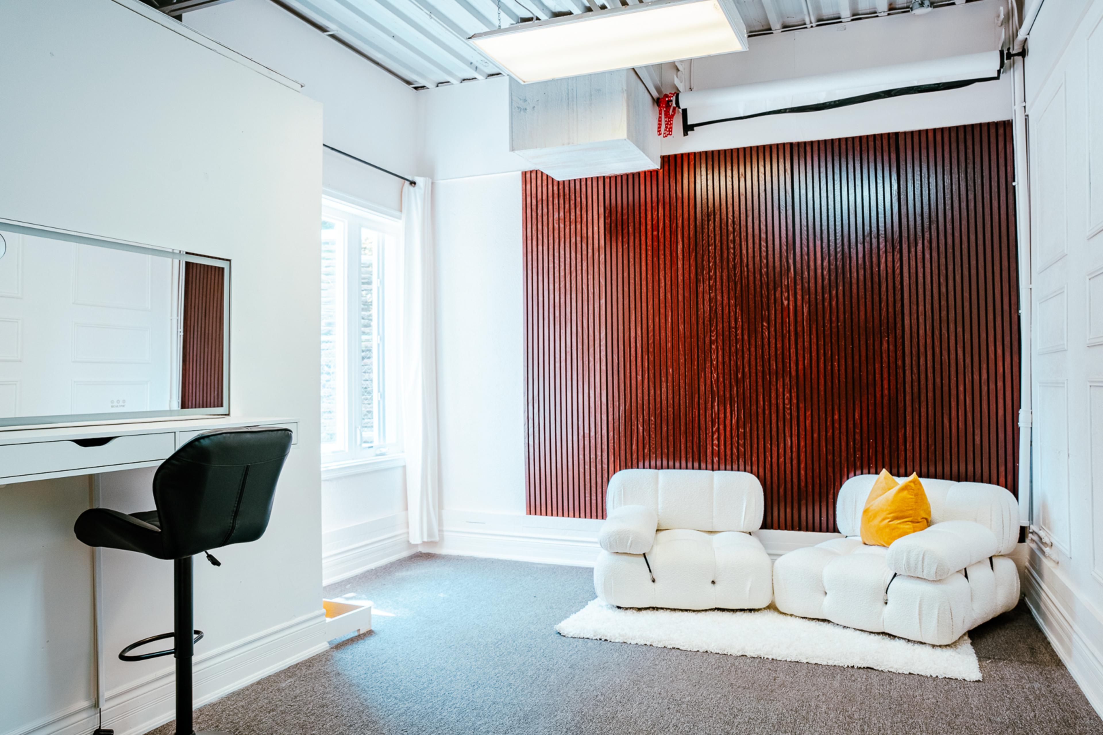 The image shows a bright room with a mirrored desk and a pair of white, textured lounge chairs against a wooden accent wall.