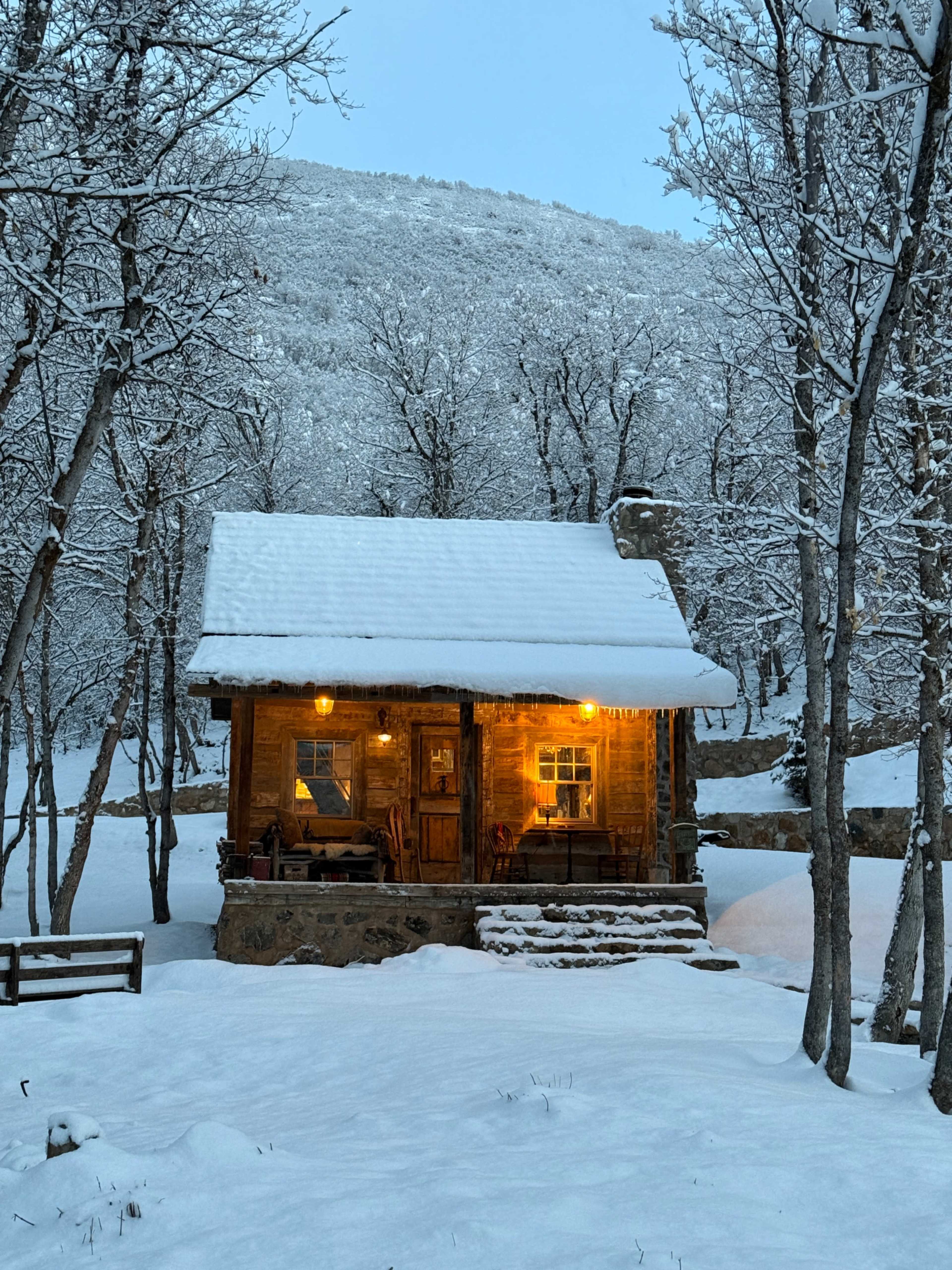 A wooden cabin sits in a snowy landscape, surrounded by bare trees and a snow-covered ground.