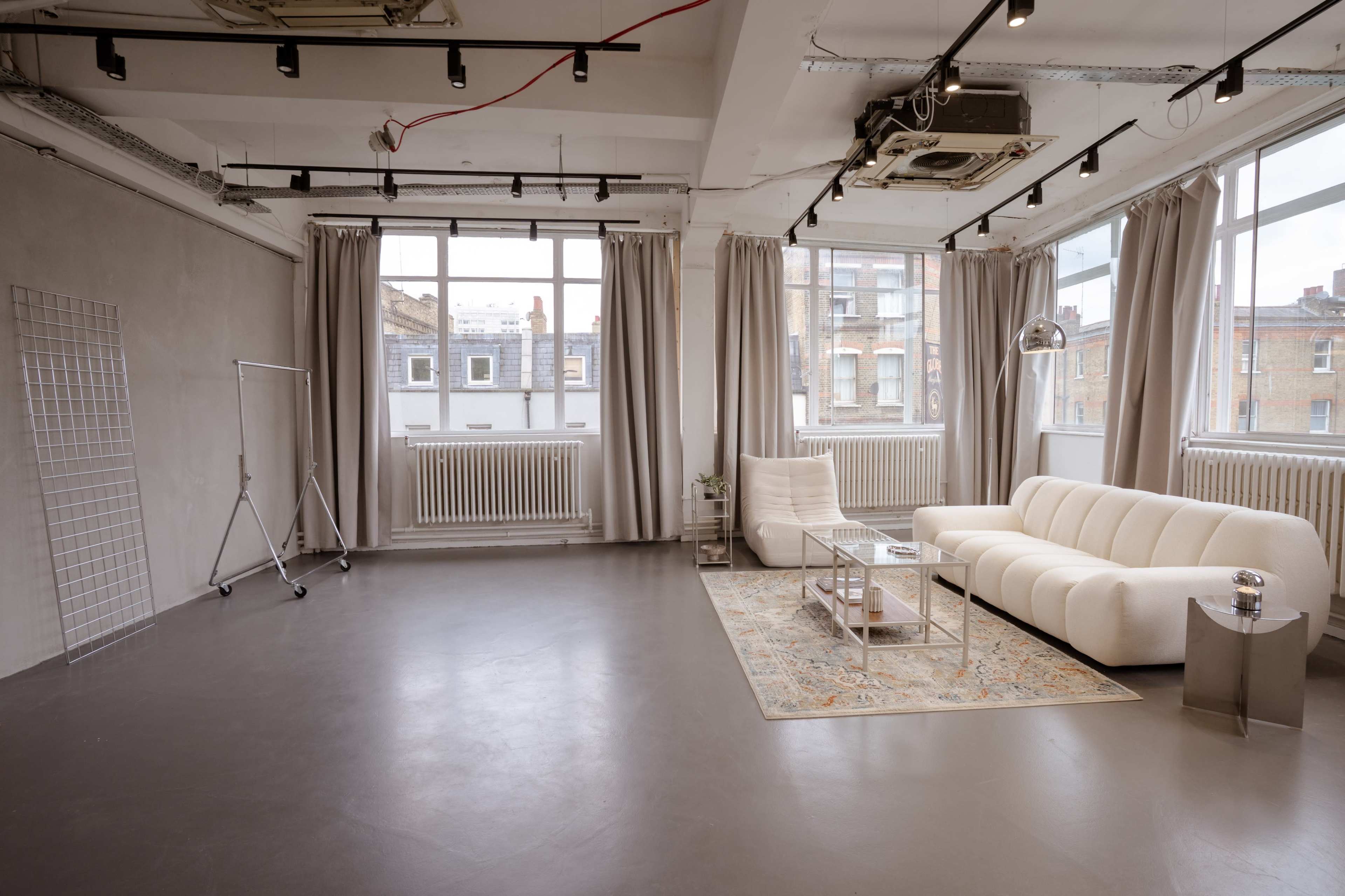 The image shows a spacious, well-lit interior featuring a white sofa, a decorative area rug, large windows, and gray walls.