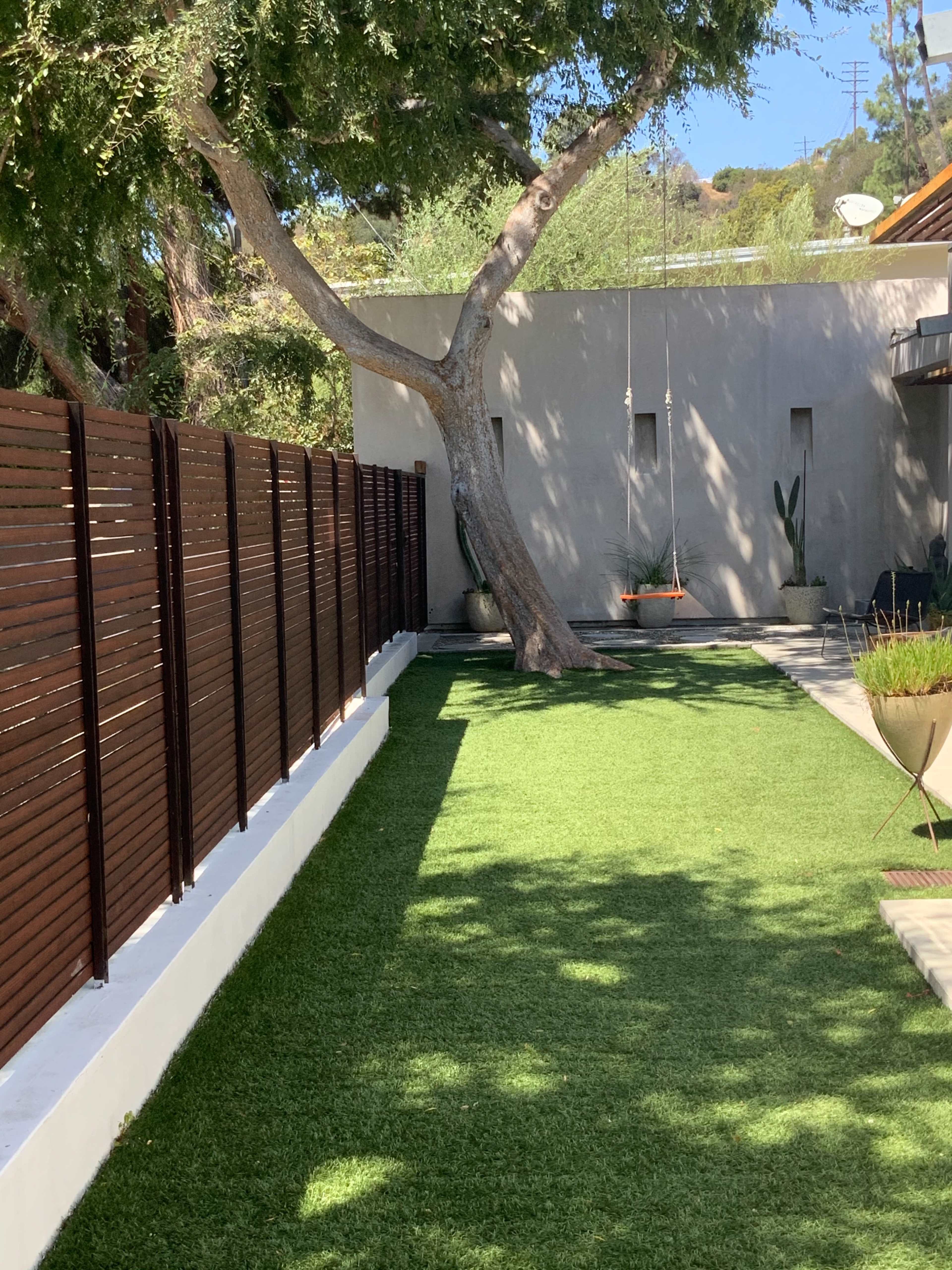 The image shows a landscaped area with artificial grass, a wooden fence, a tree, and a wall adorned with a hanging planter.