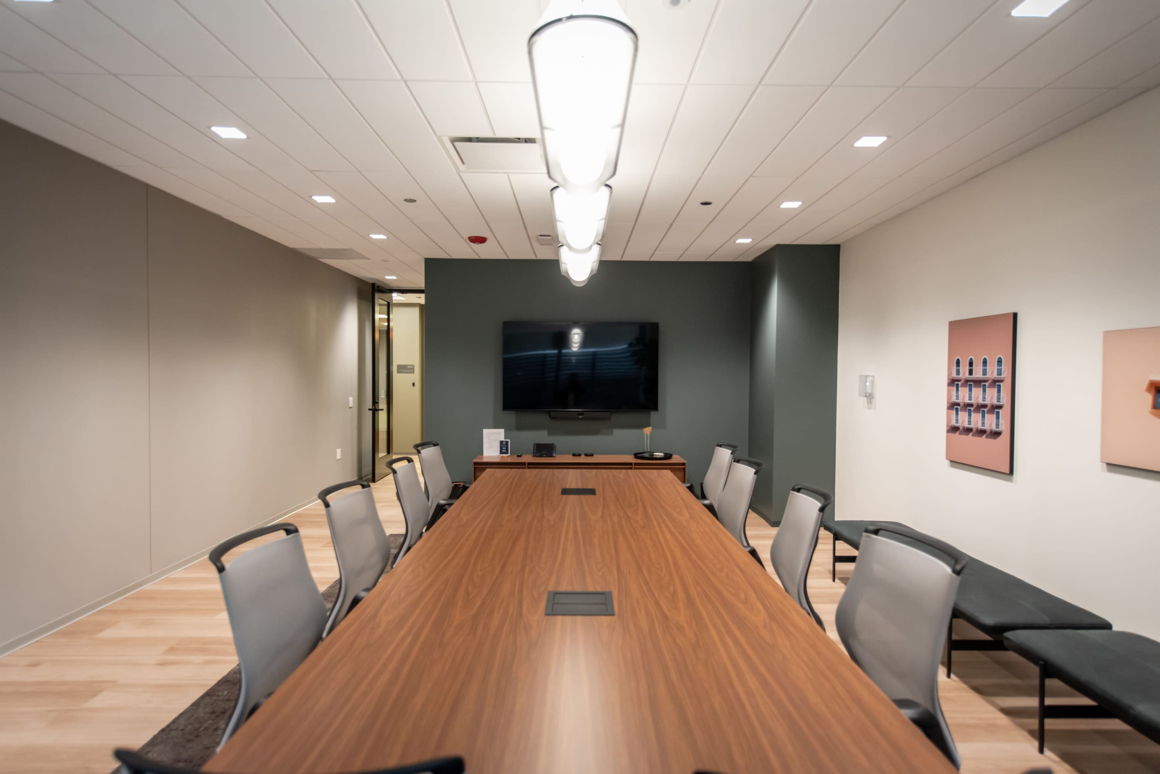 A large conference room features a long wooden table surrounded by gray chairs, with a wall-mounted TV and art displayed on the walls.