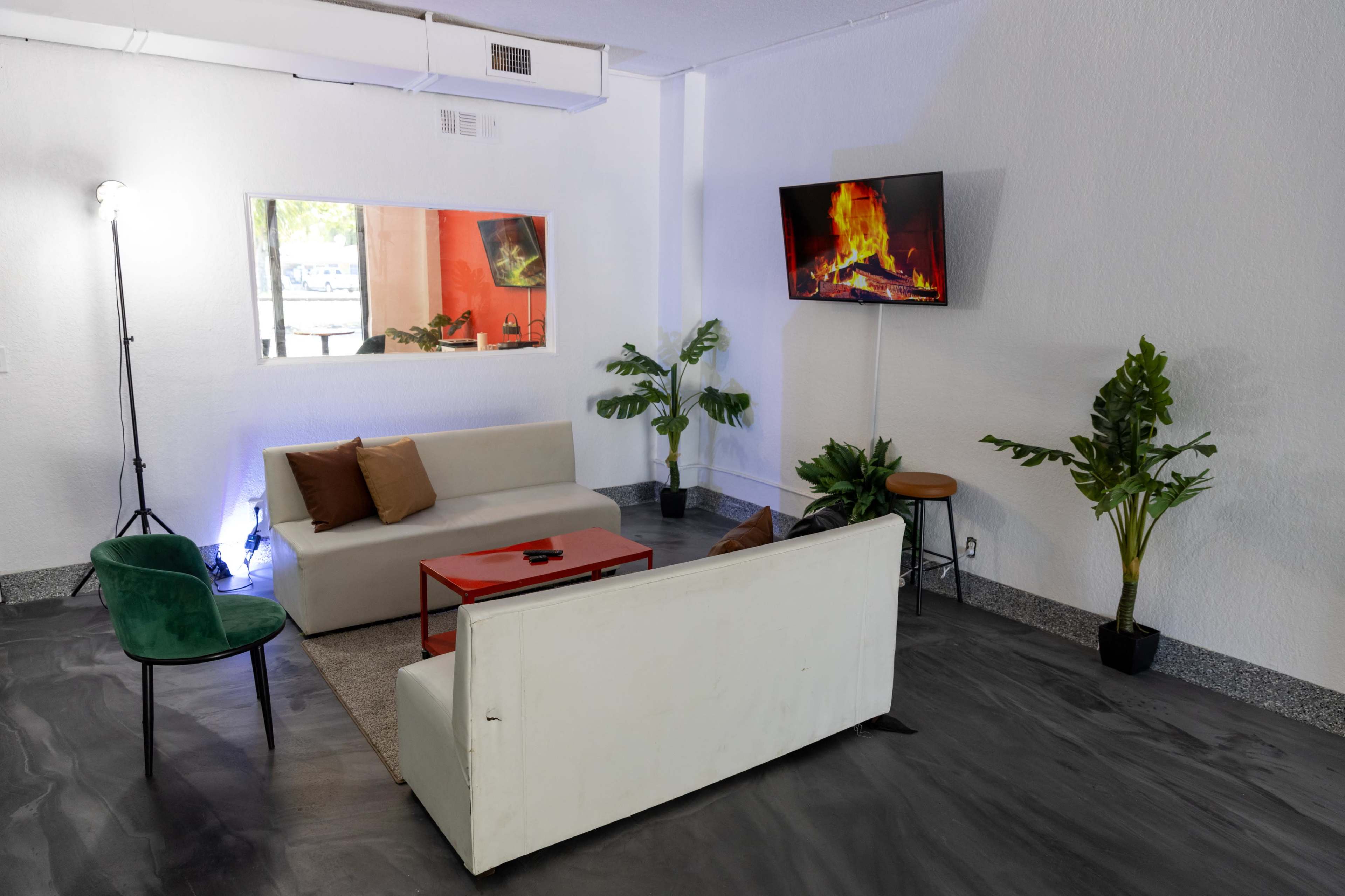 A modern living room features a neutral sofa, a red coffee table, a television displaying a fireplace, and potted plants.