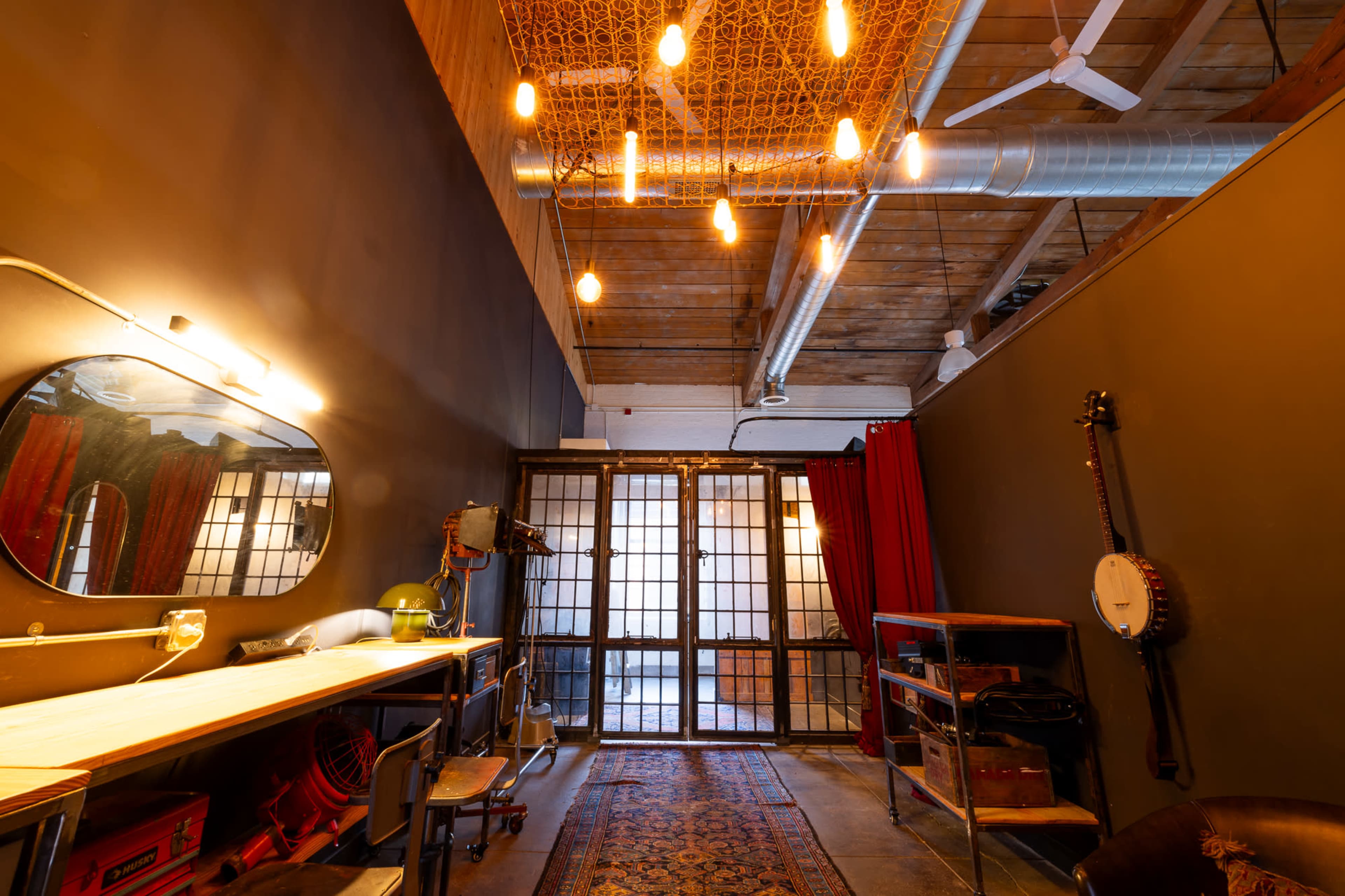 A loft-style room with high wooden ceilings, a long table with a mirror, various light fixtures, and a doorway flanked by red curtains.