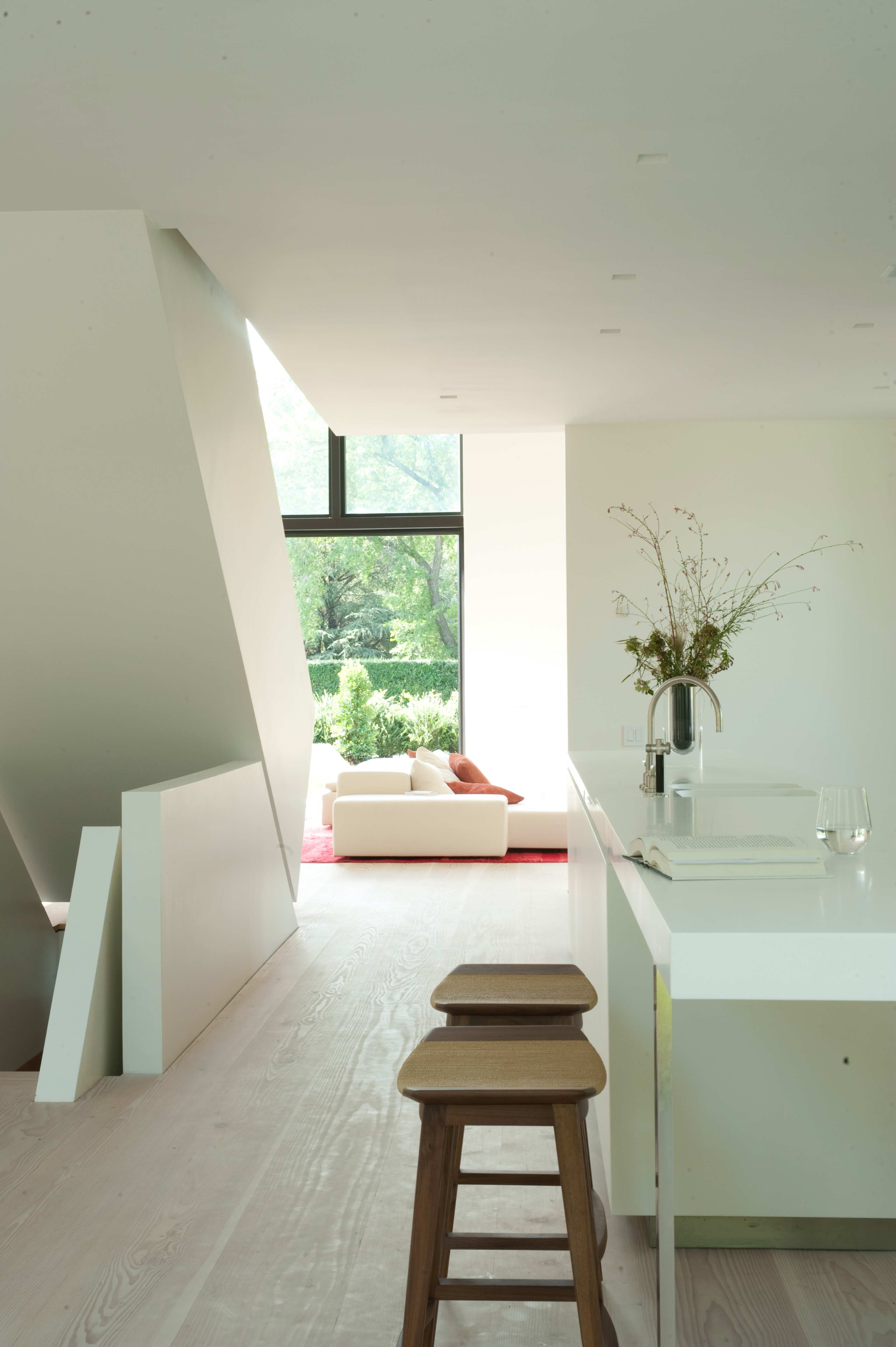 A modern, minimalist kitchen with white walls and cabinetry features wooden stools and a view of a bright living area through large windows.
