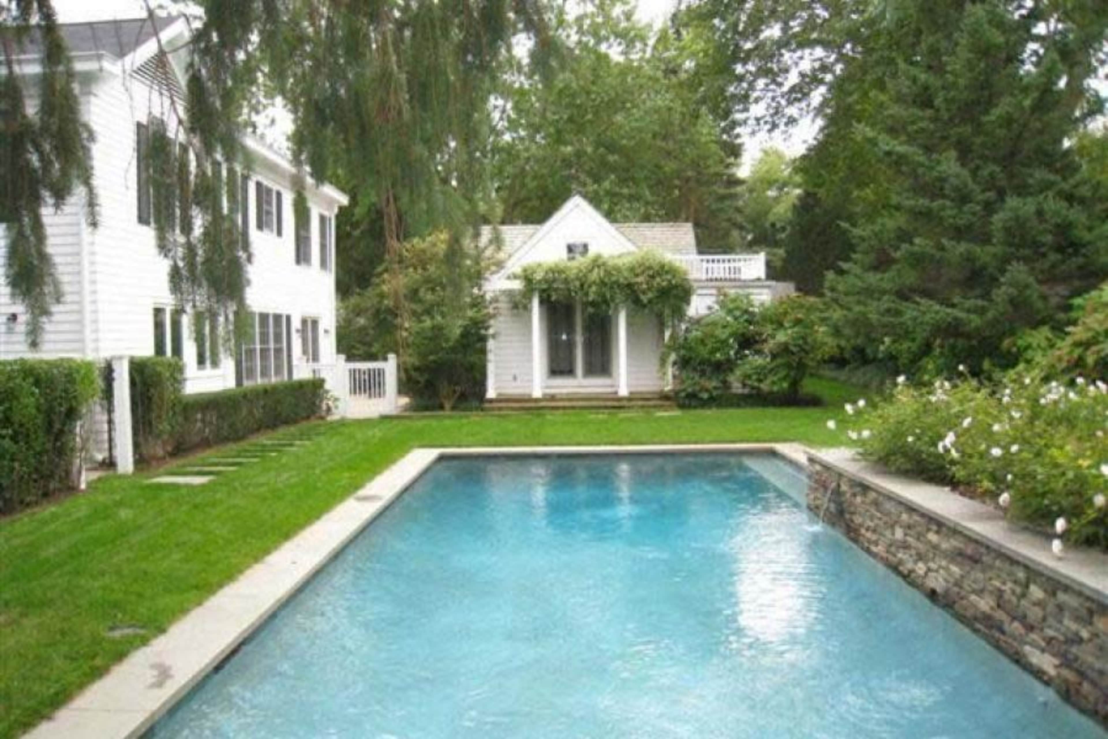 A well-maintained backyard features a clear swimming pool surrounded by greenery and a small white cottage in the background.