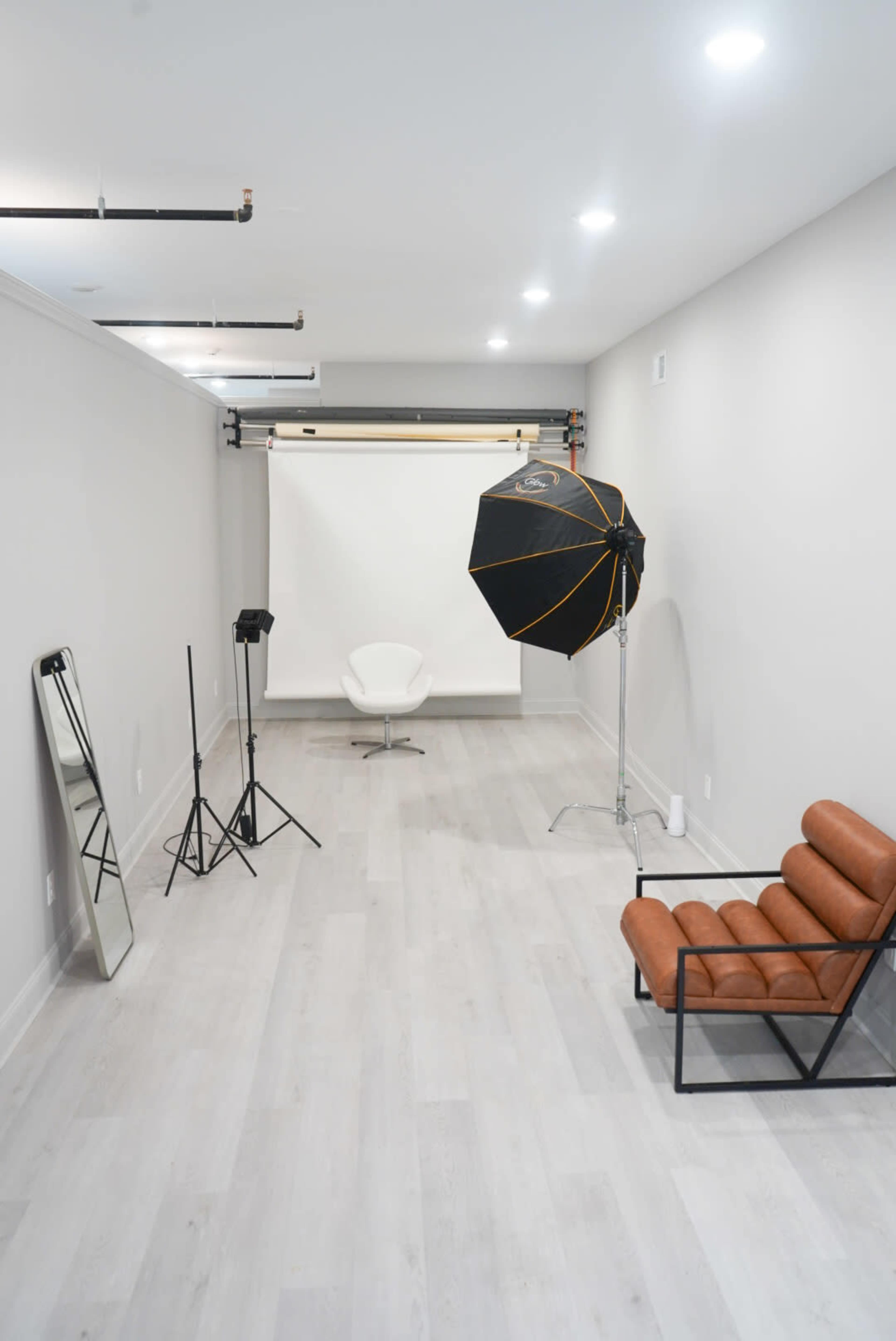 The image shows a minimalist studio space featuring a white backdrop, a single chair, a large mirror, a photography light, and a brown leather sofa.
