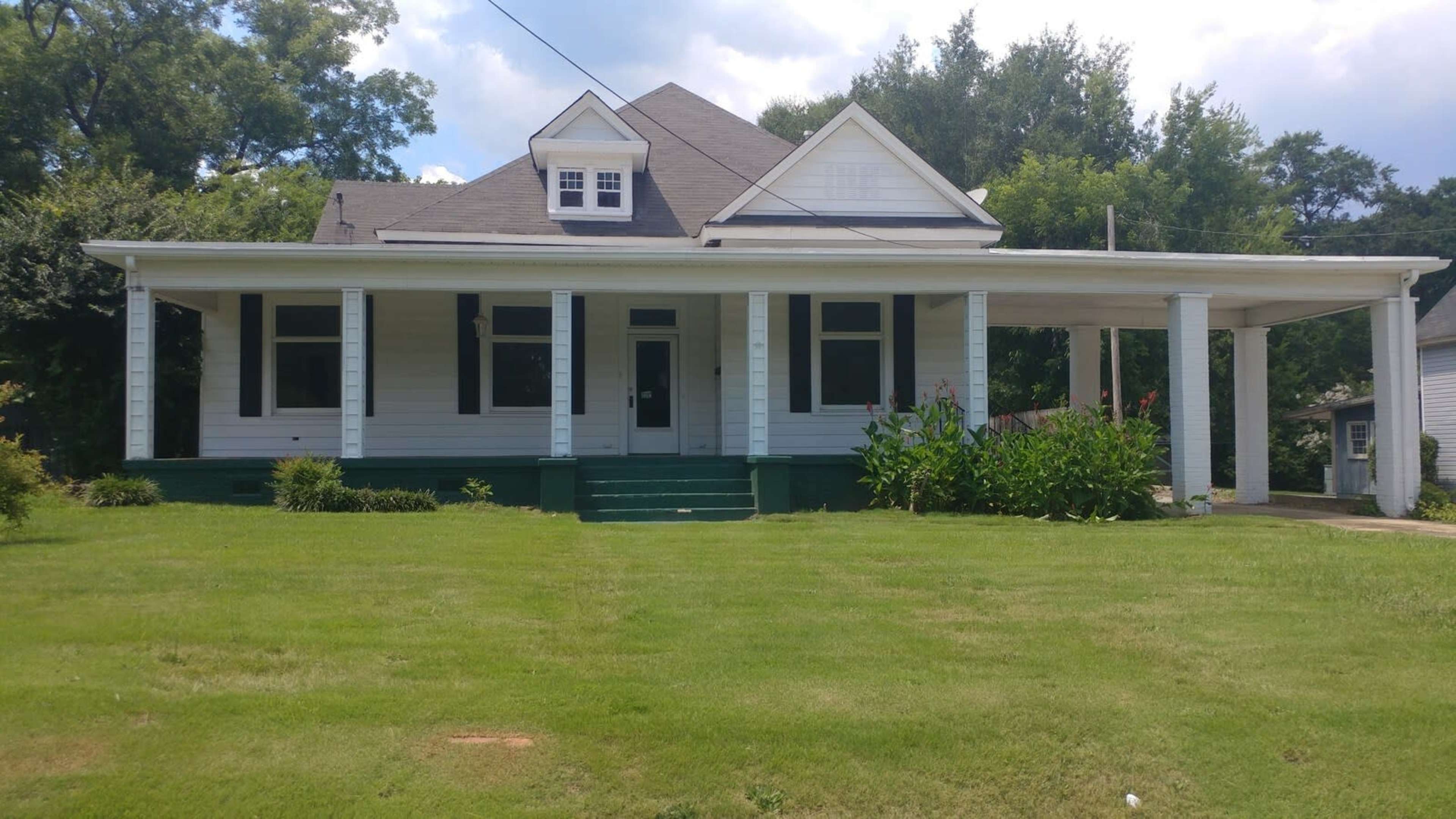 A two-story white house with a covered porch and green steps is surrounded by grass and shrubs.
