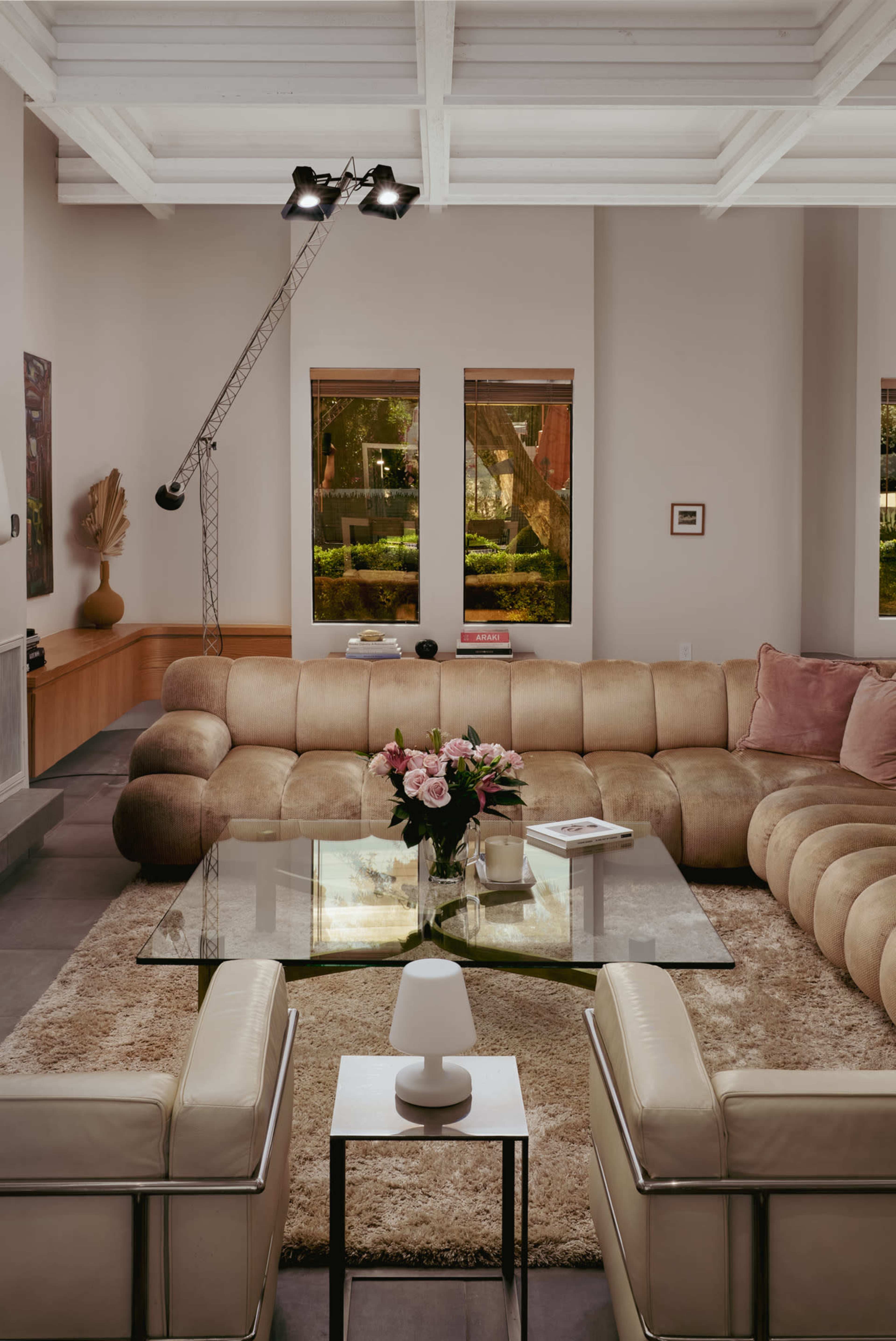 The spacious living room features a large beige sectional sofa, a glass coffee table, and a stylish lamp, with ample natural light streaming in through the windows.