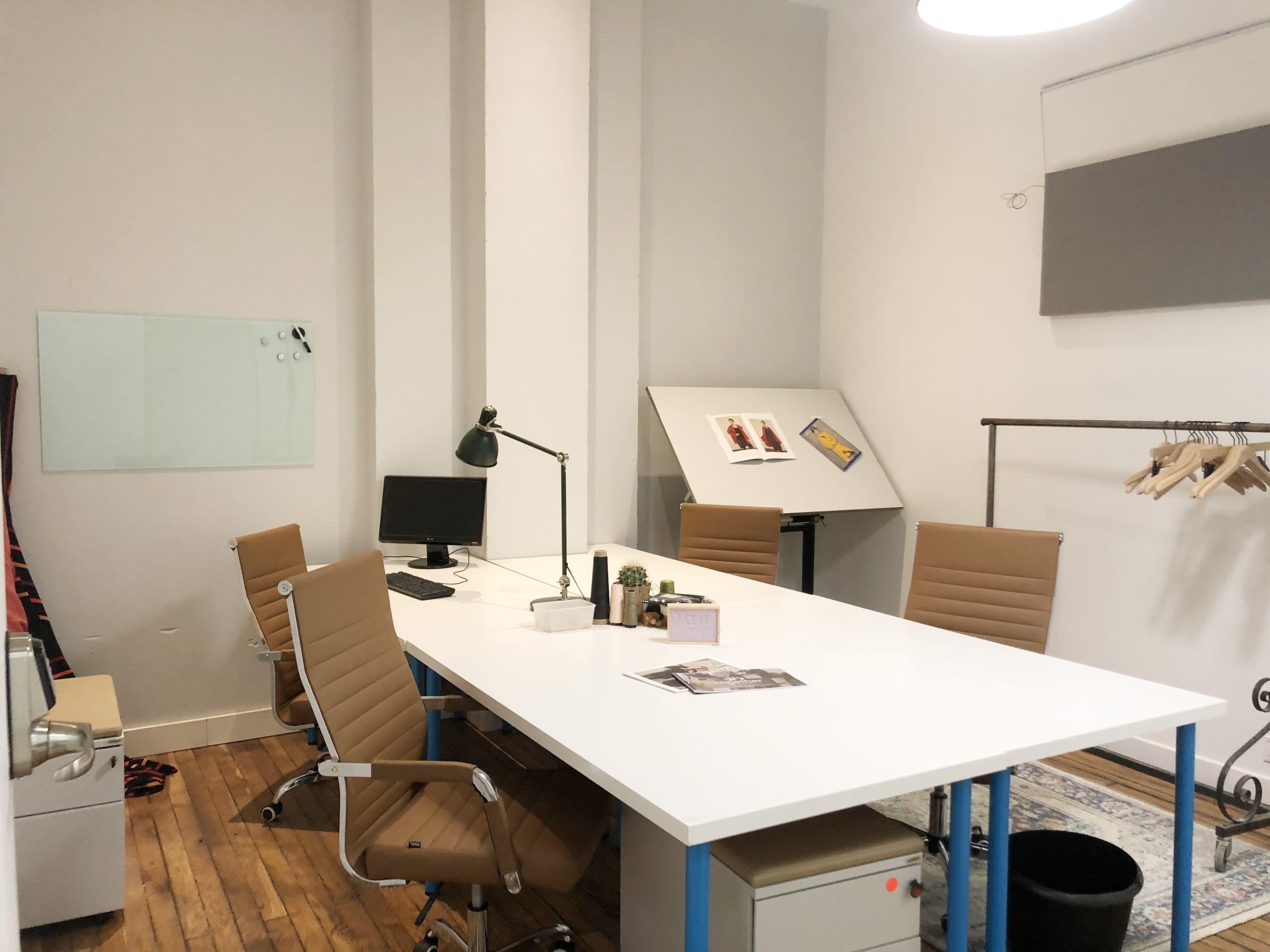 The image shows a modern office space featuring a long white table with brown chairs, a computer, a floor lamp, and a display board on an easel.