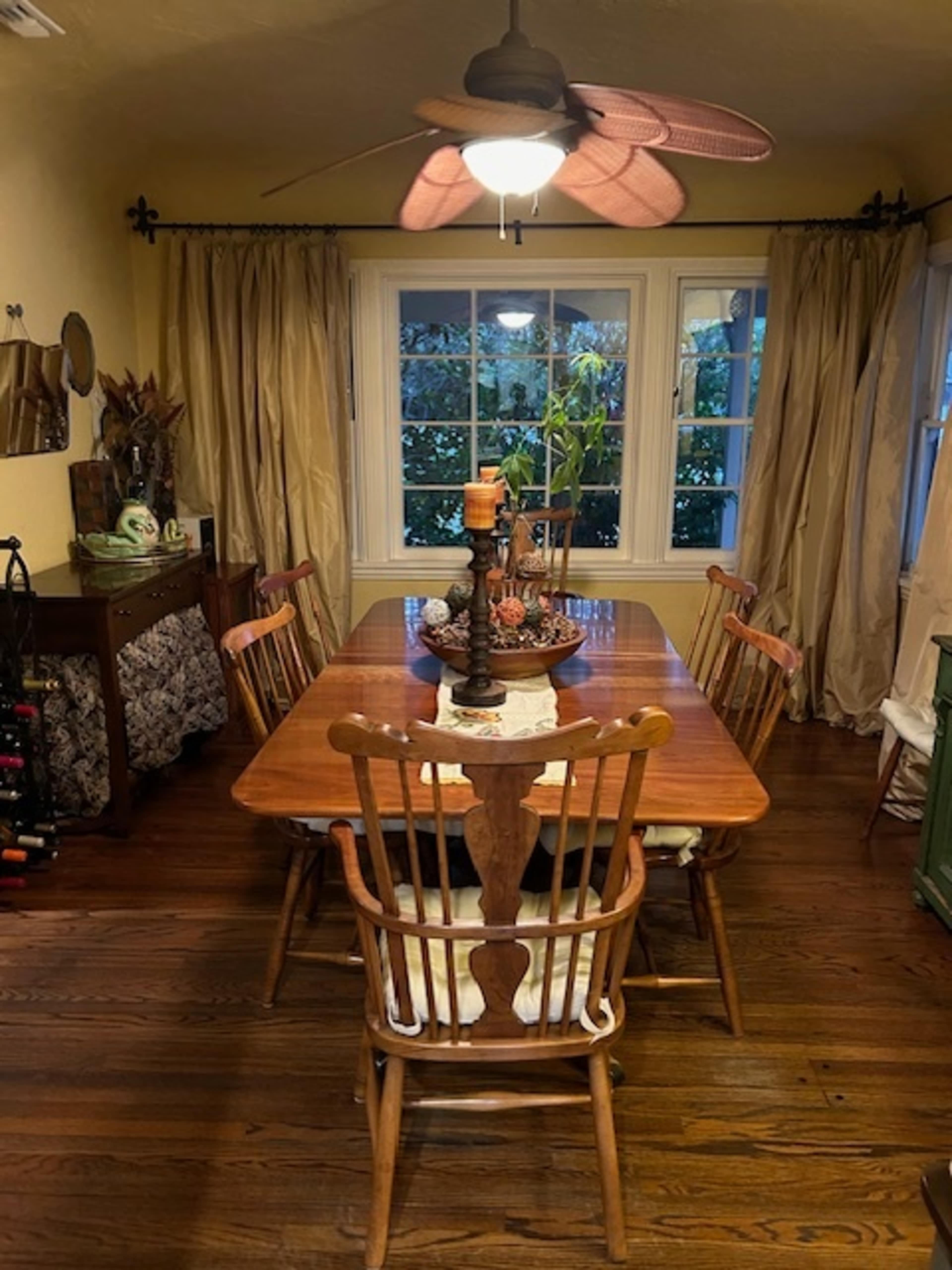 A wooden dining table is surrounded by chairs in a warmly lit room with large windows and curtains.