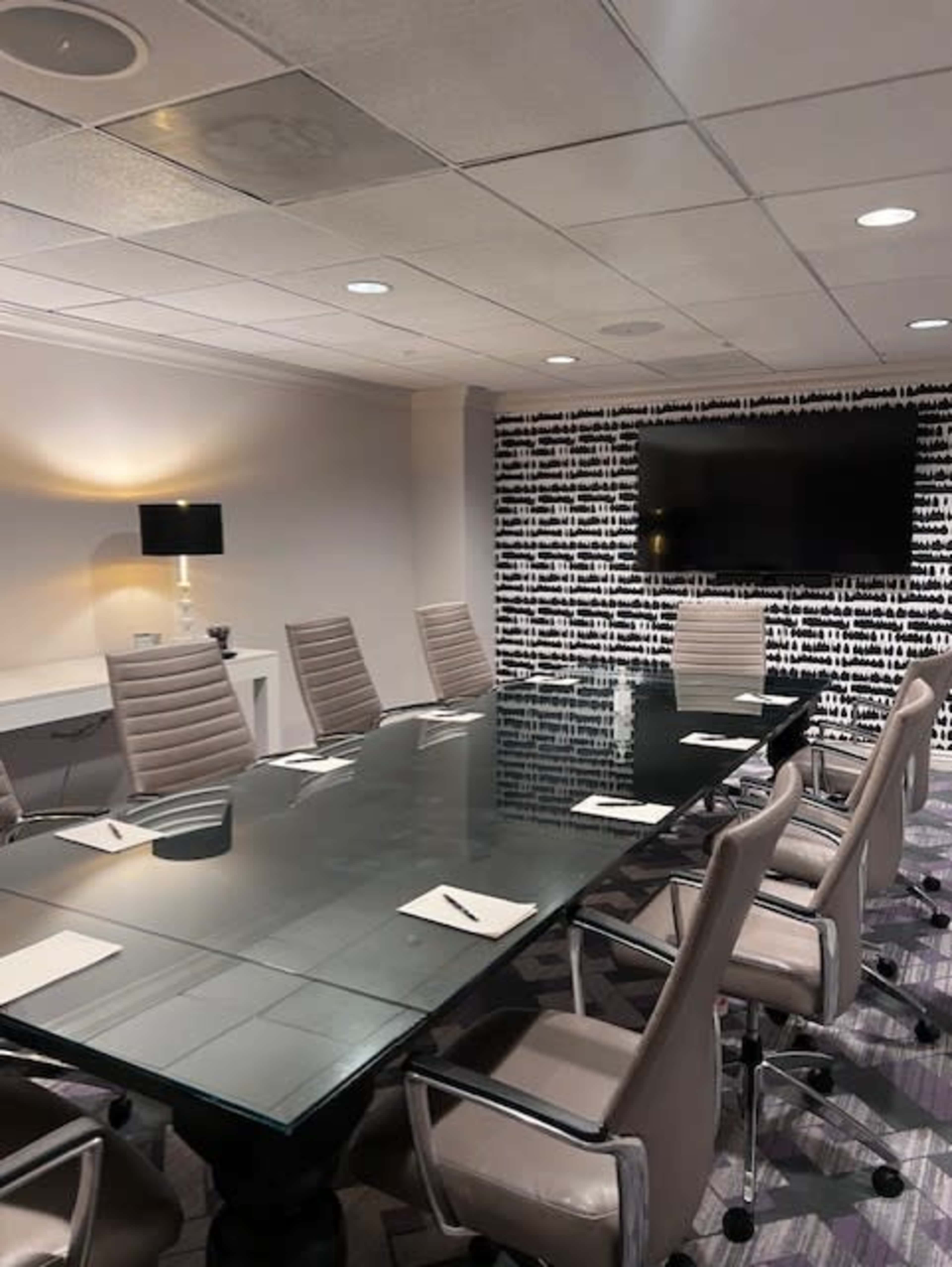 A modern conference room features a long glass table surrounded by eight ergonomic chairs, with a wall-mounted screen and a patterned backdrop.