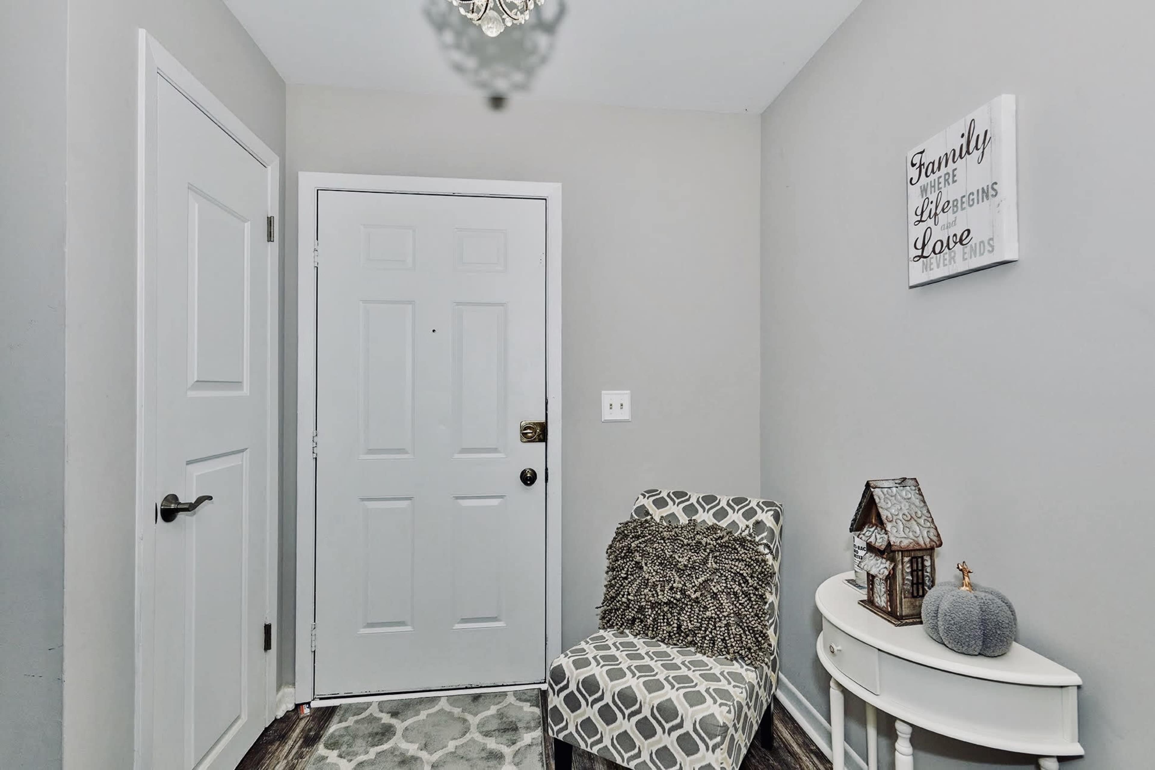 The image shows a small entryway with a gray wall, a white door, a patterned chair, and a decorative table holding a lantern and a decorative item.