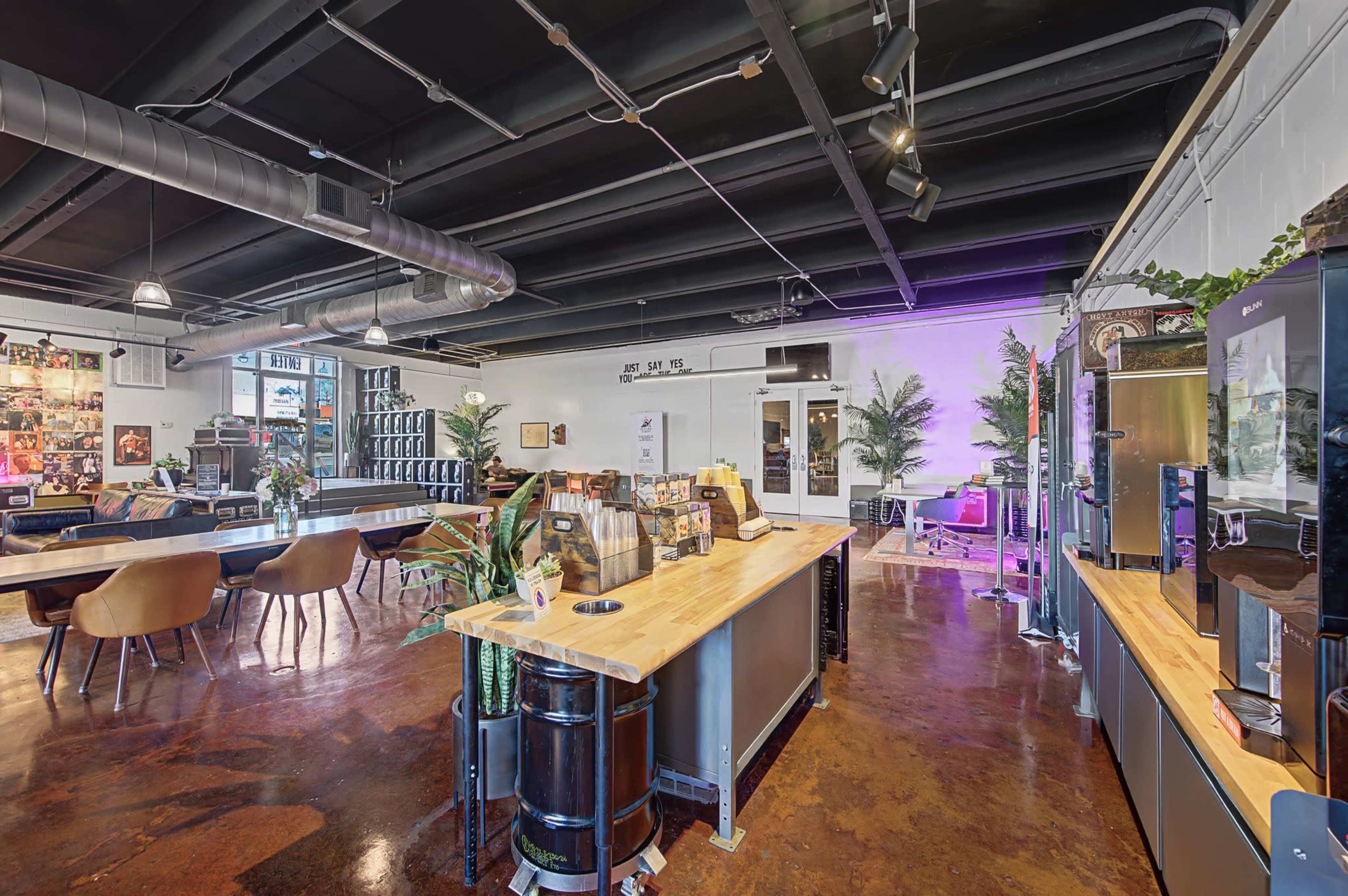 The image shows a modern, open workspace with a bar-counter area, seating, and decorative plants.