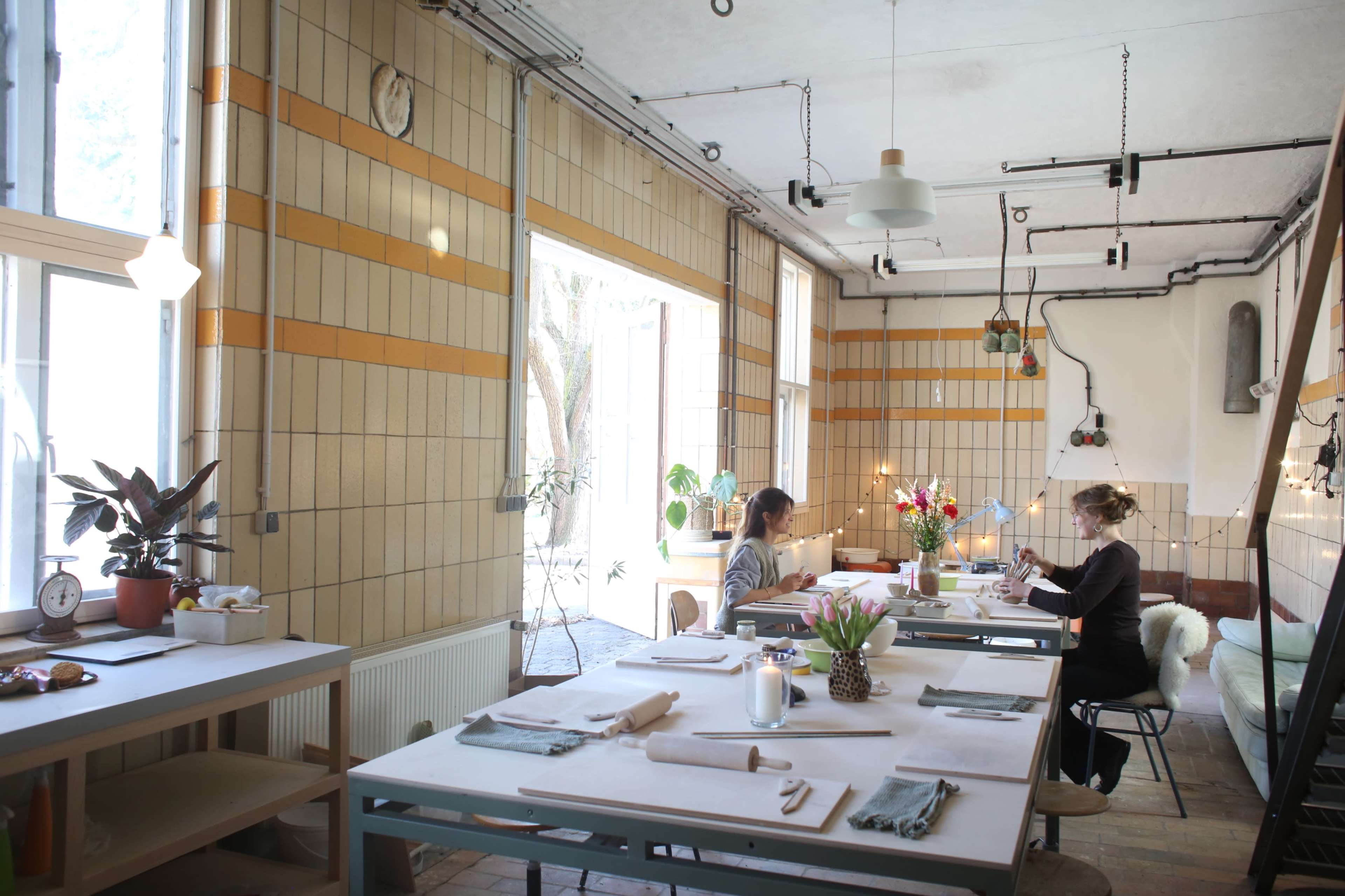 The image shows a bright, minimalist dining area with two women seated at separate tables, surrounded by plants and large windows.