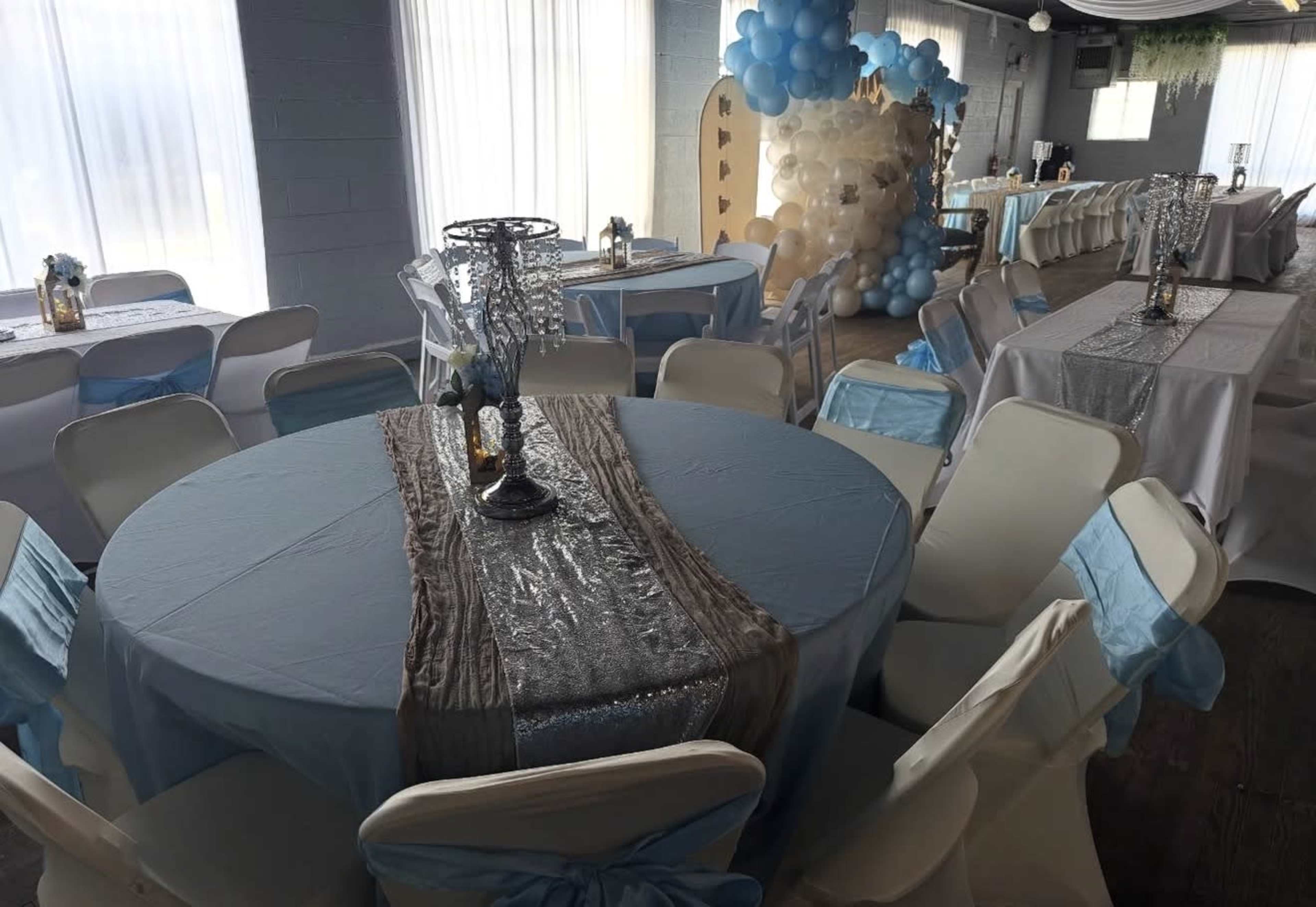 The image shows a decorated indoor event space with round tables covered in light blue tablecloths, white chairs with blue sashes, and a backdrop of balloons.