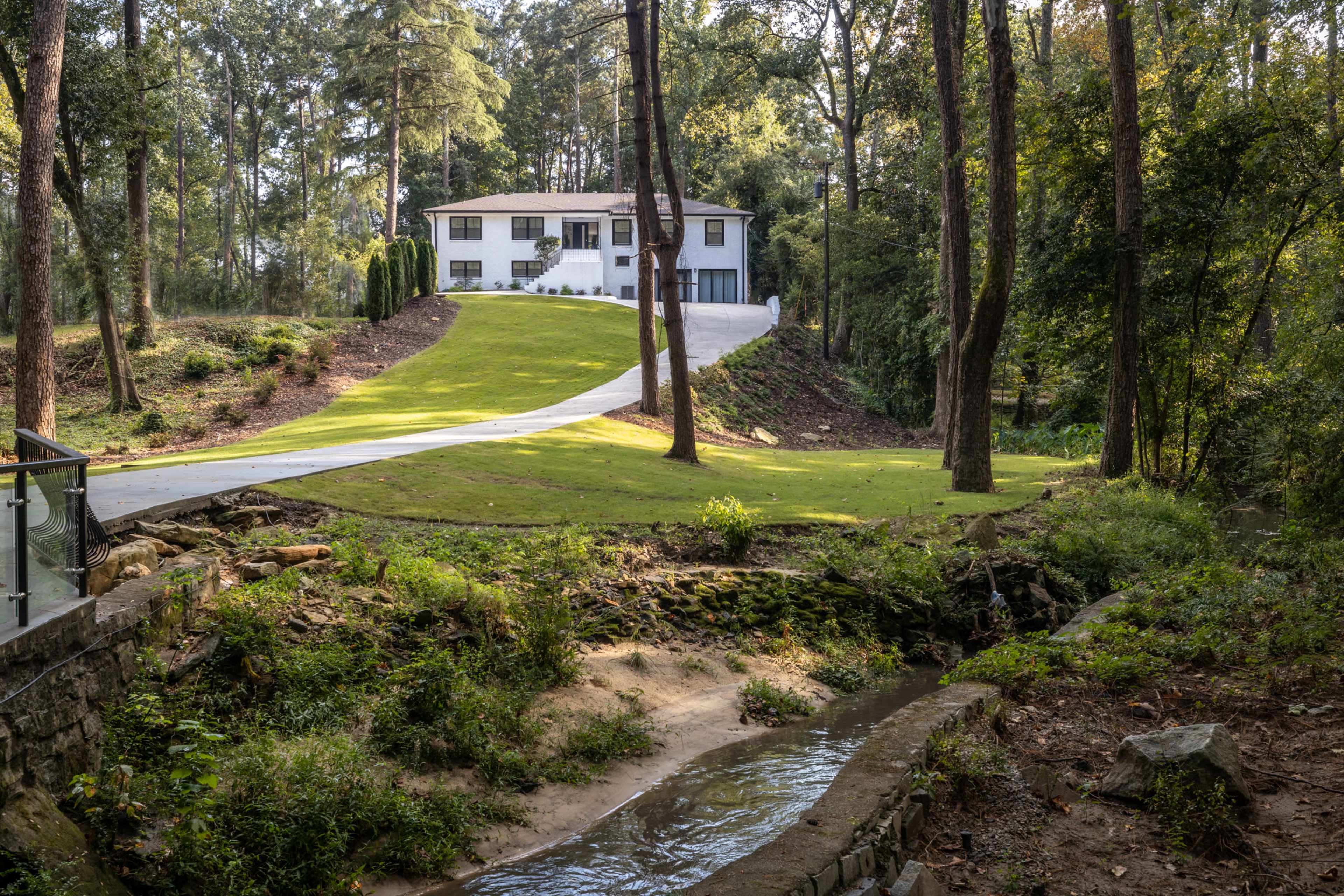A modern two-story house sits on a green hillside, bordered by tall trees, with a paved driveway leading to its entrance.