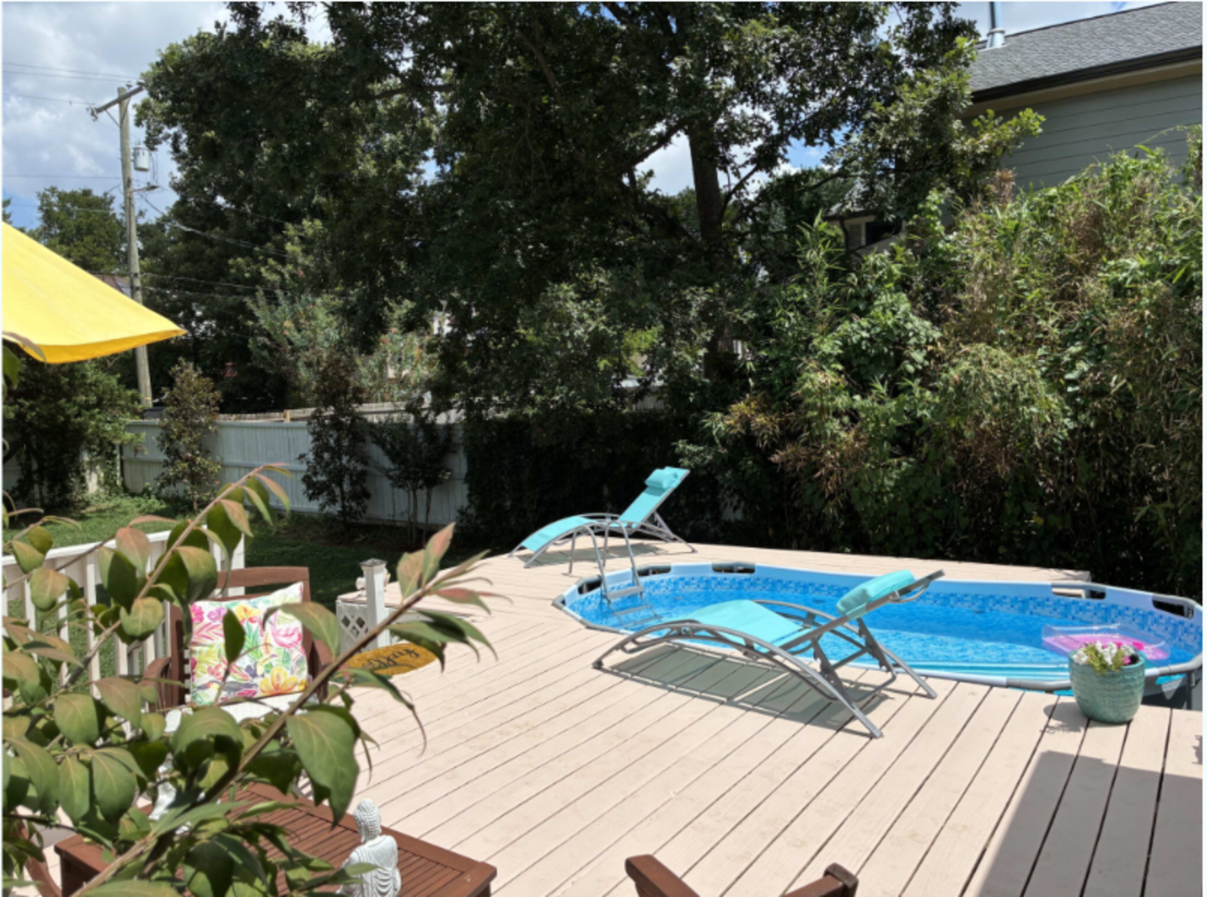 The scene features a pool surrounded by a wooden deck, with lounge chairs positioned nearby and greenery in the background.