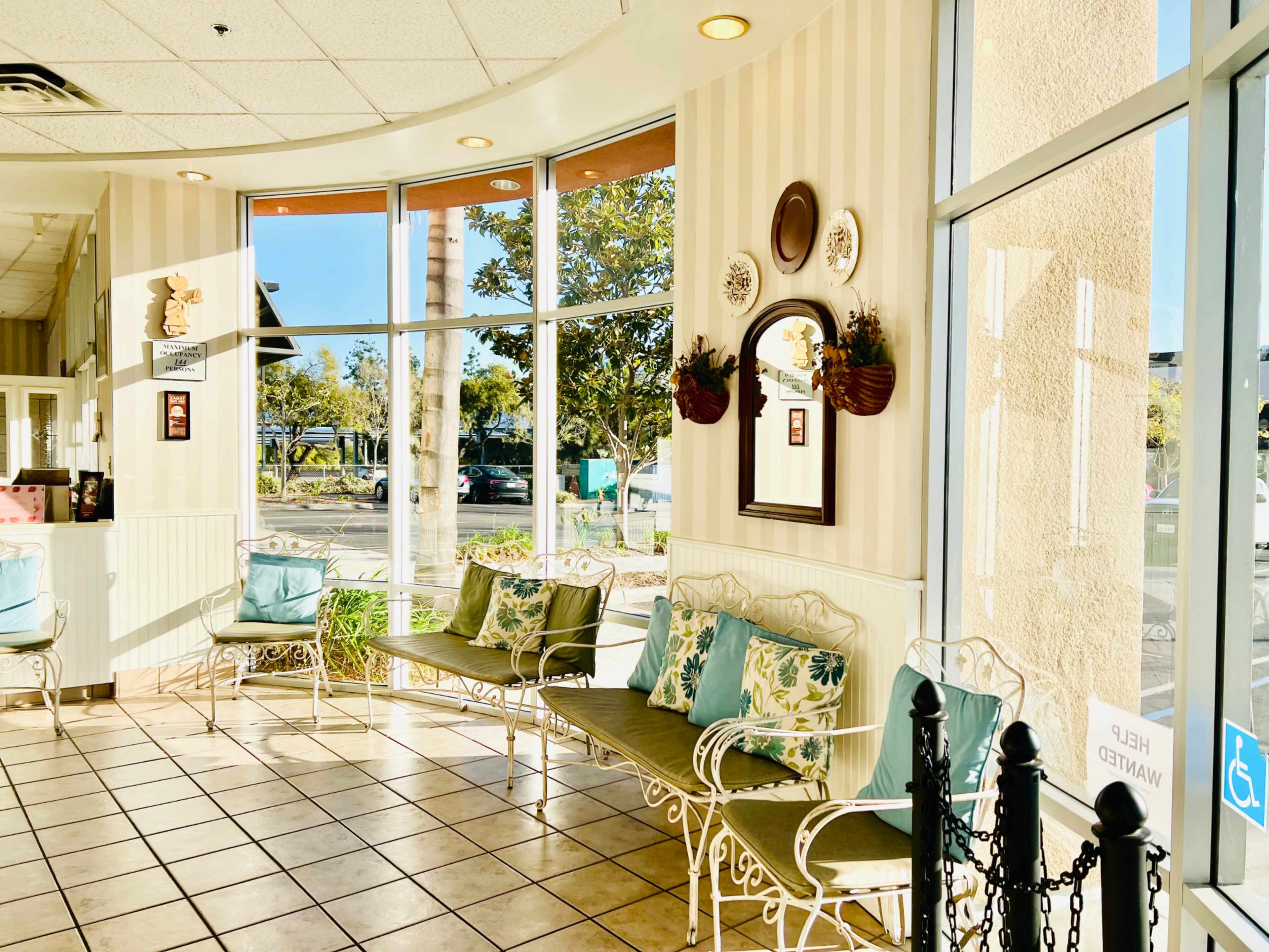 The image shows a waiting area with metal benches adorned with cushions, large windows letting in natural light, and decorative elements on the walls.