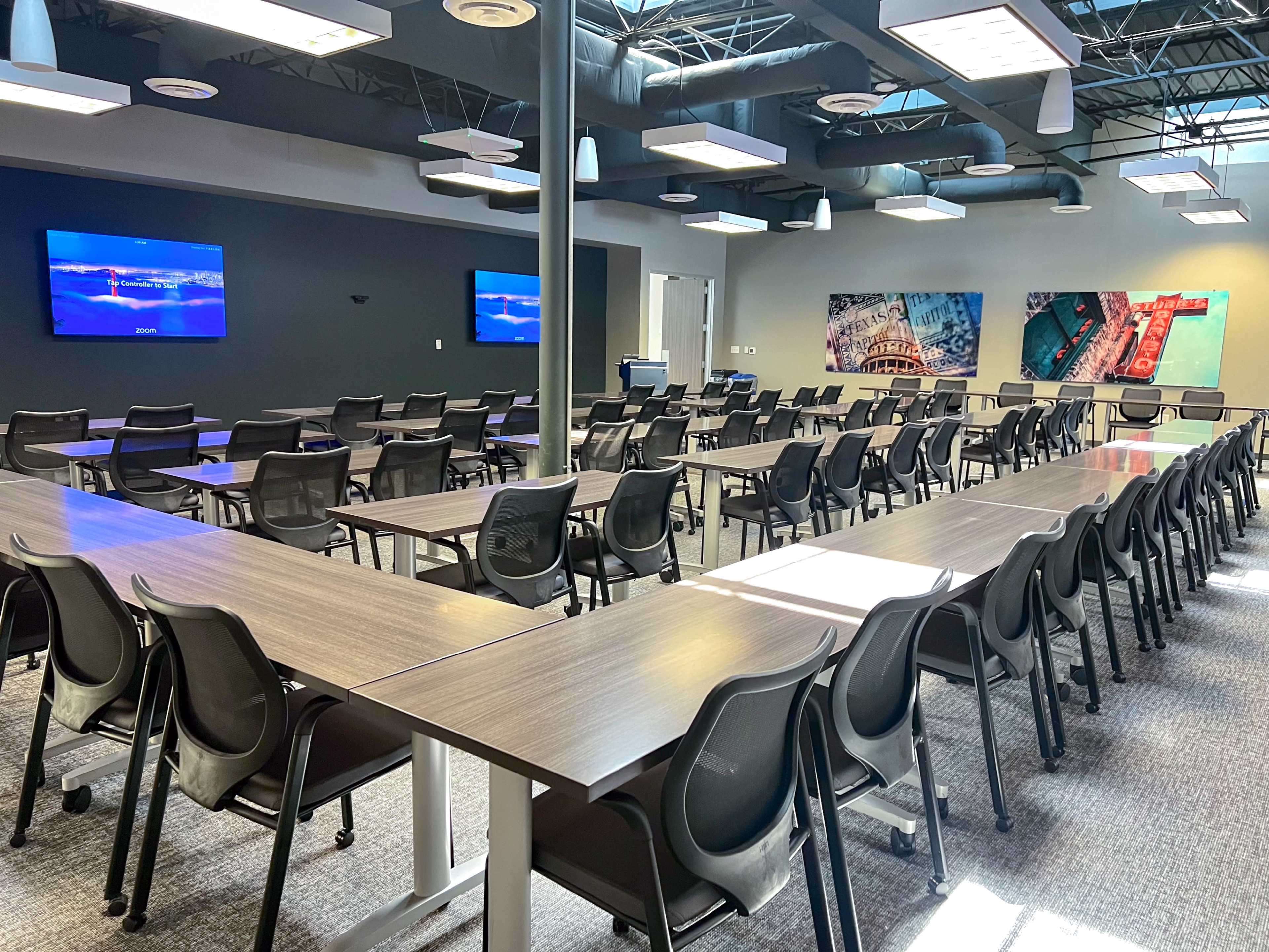 The image shows a spacious meeting room with multiple rows of tables and chairs, two screens mounted on the front wall, and large windows allowing natural light.