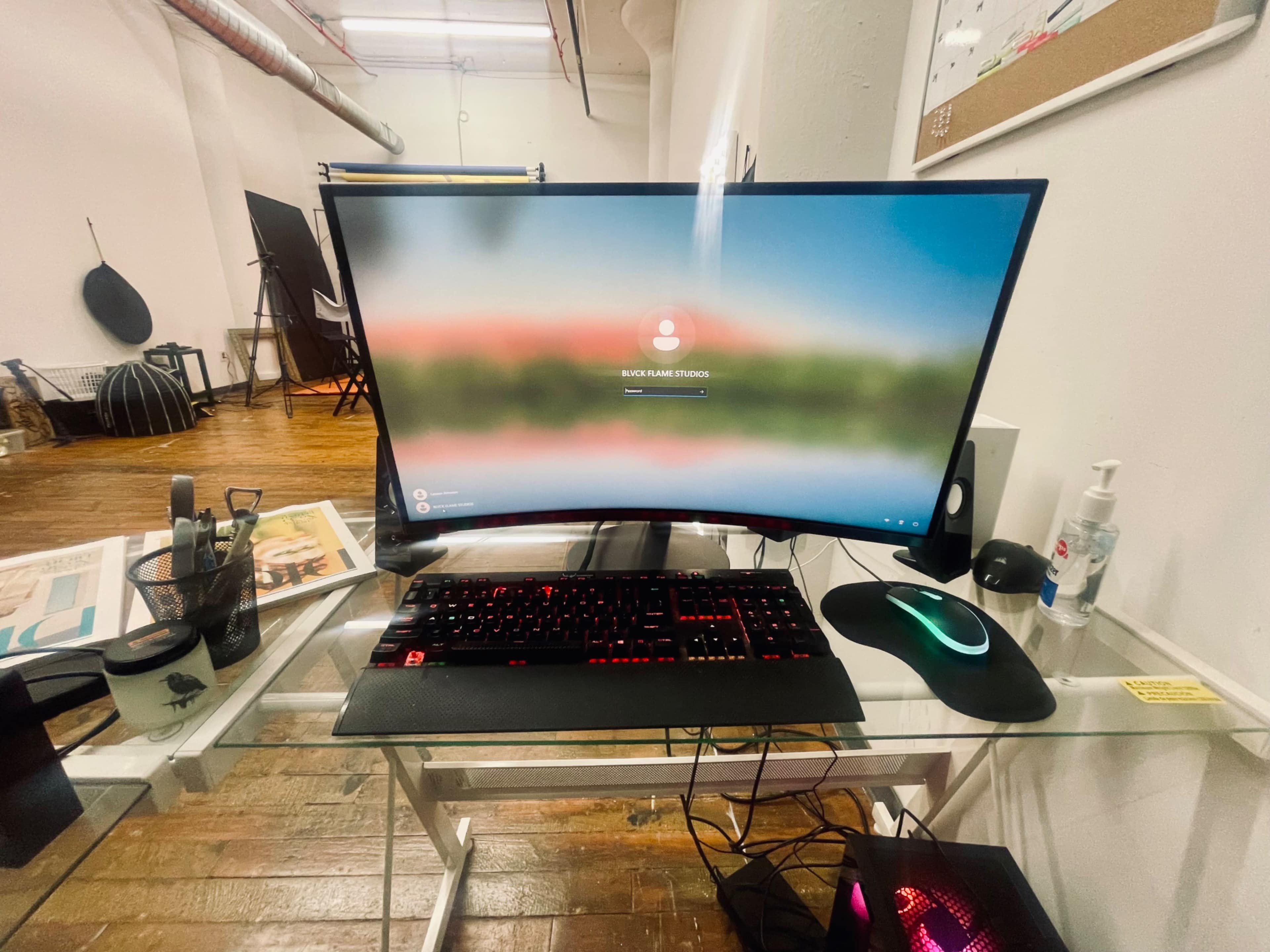 A curved computer monitor displaying a login screen is positioned on a glass desk in a brightly lit room with wooden flooring.