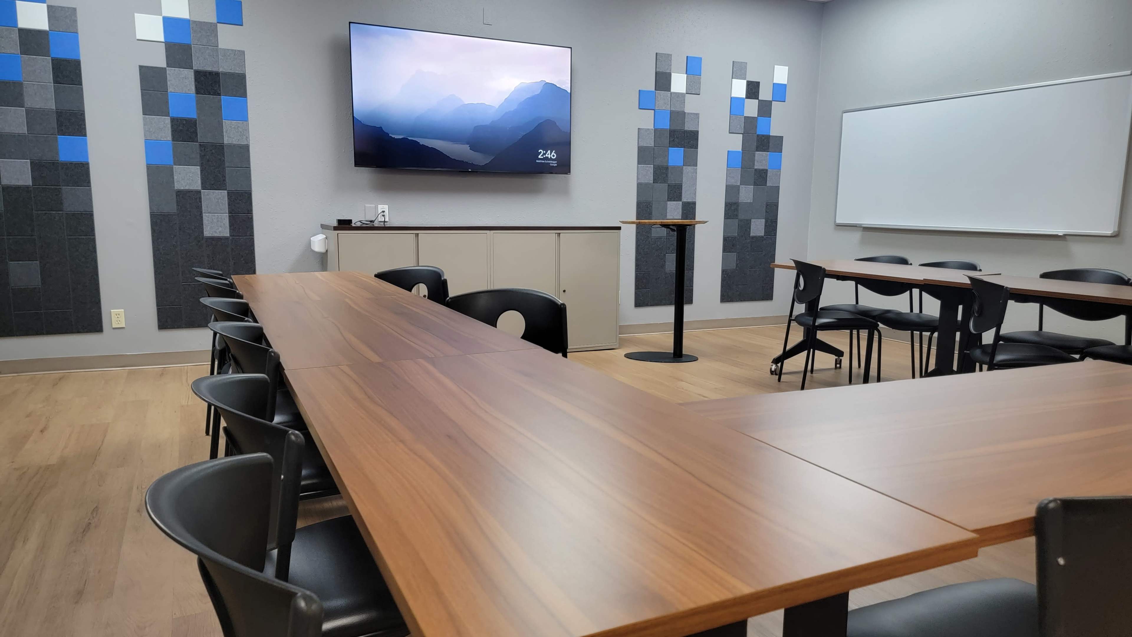 The image shows a modern conference room with a long wooden table, several black chairs, a wall-mounted television, and a whiteboard.