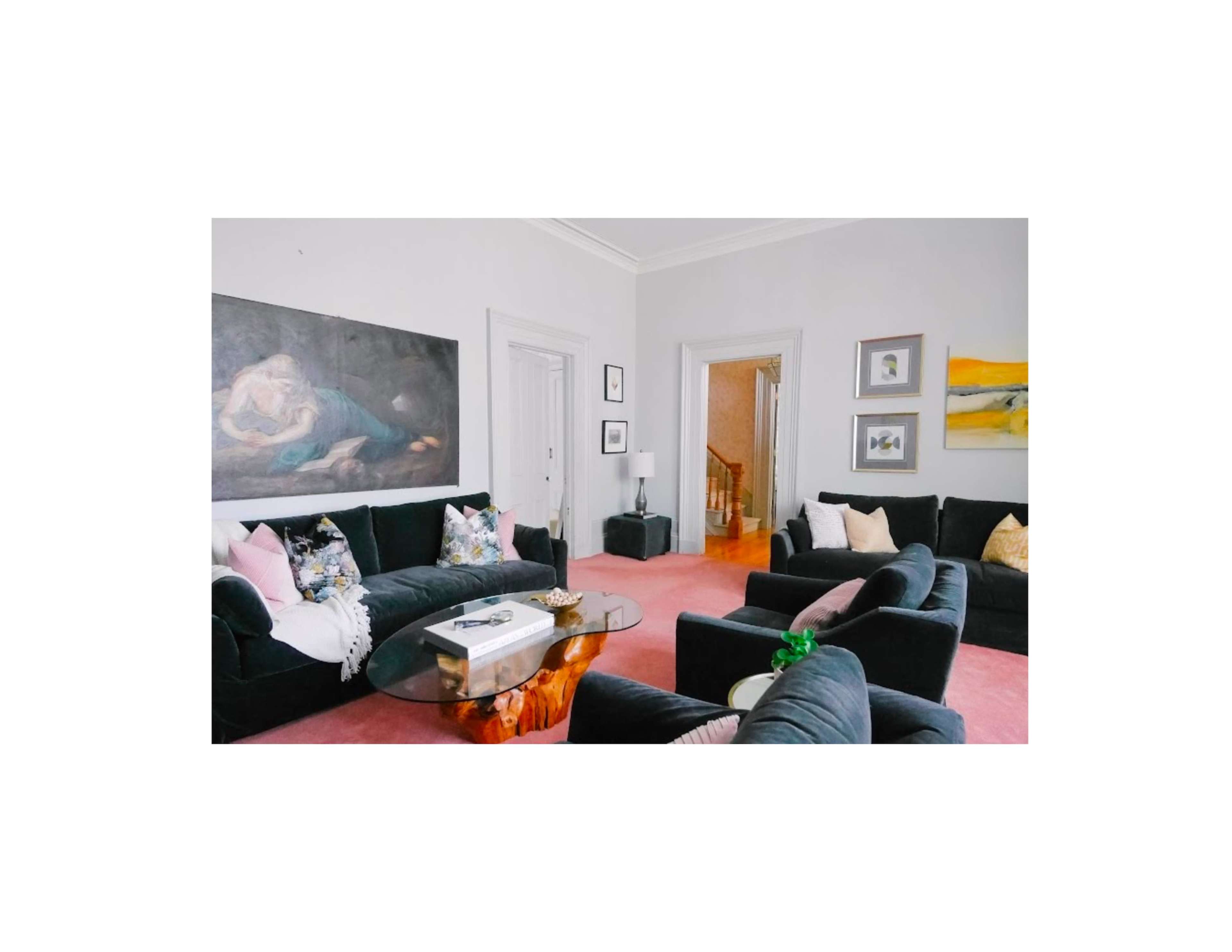 The image shows a living room with black sofas, a glass coffee table, a large painting on the wall, and light-colored walls, featuring a staircase in the background.