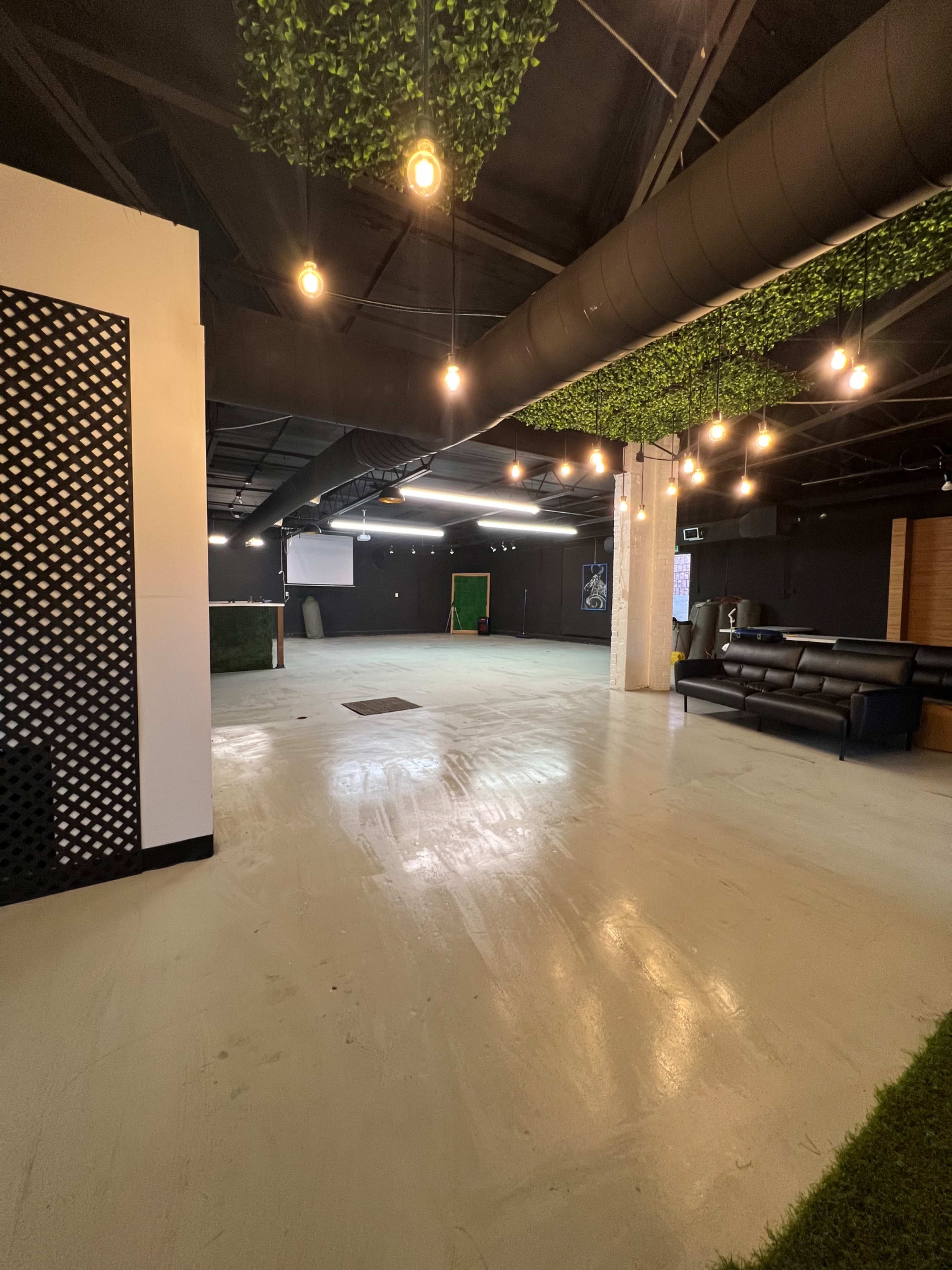 A spacious interior with a concrete floor, a green wall feature, and various seating areas illuminated by hanging lights.