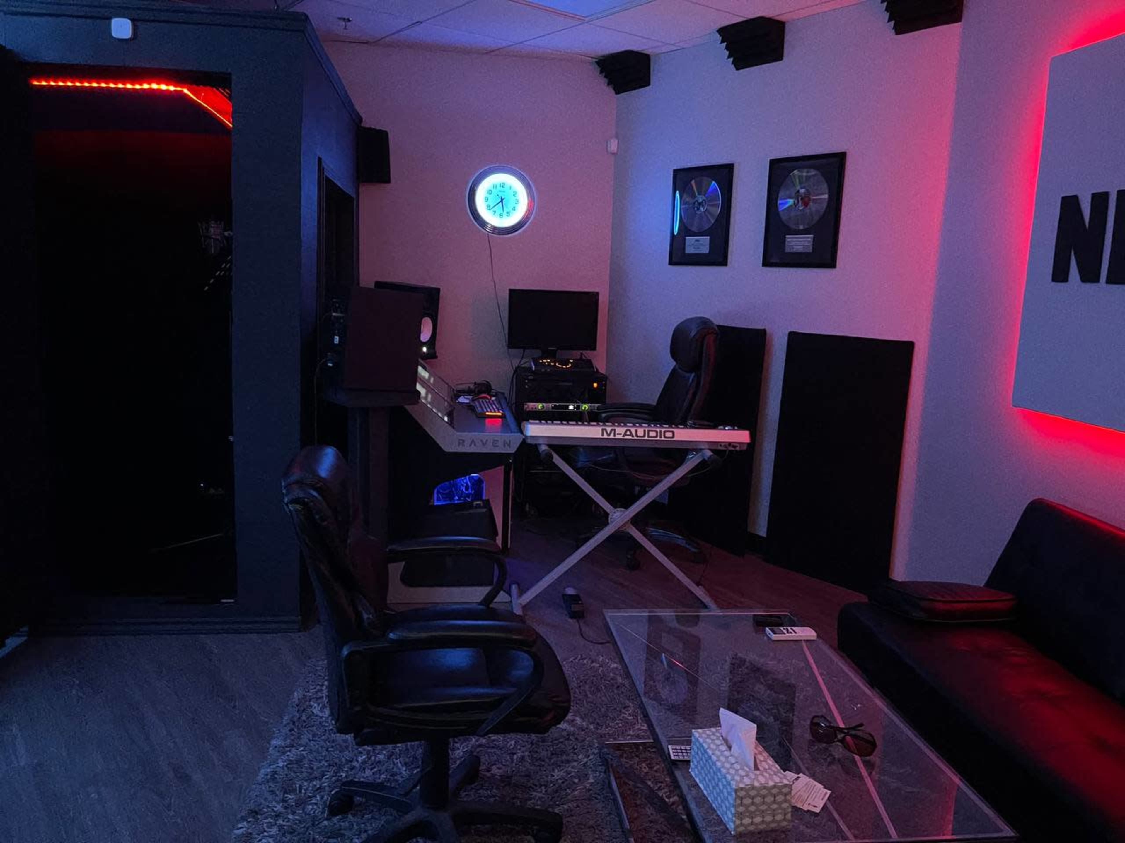 A music studio with a mixing desk, keyboard, and soundproof booth, illuminated by red and blue lighting.