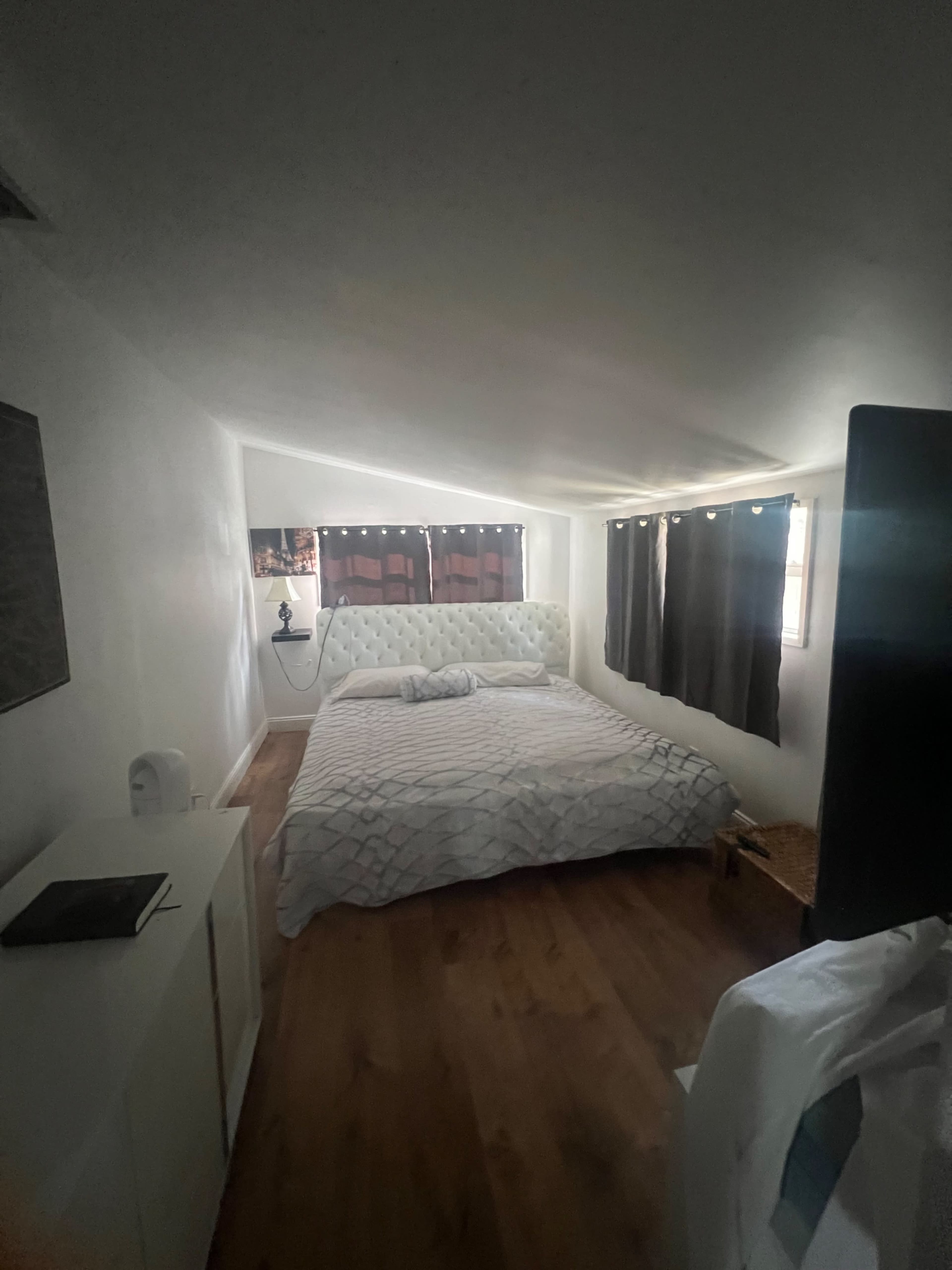 A bedroom featuring a bed with white bedding, black curtains over two windows, and a small white dresser along the wall.