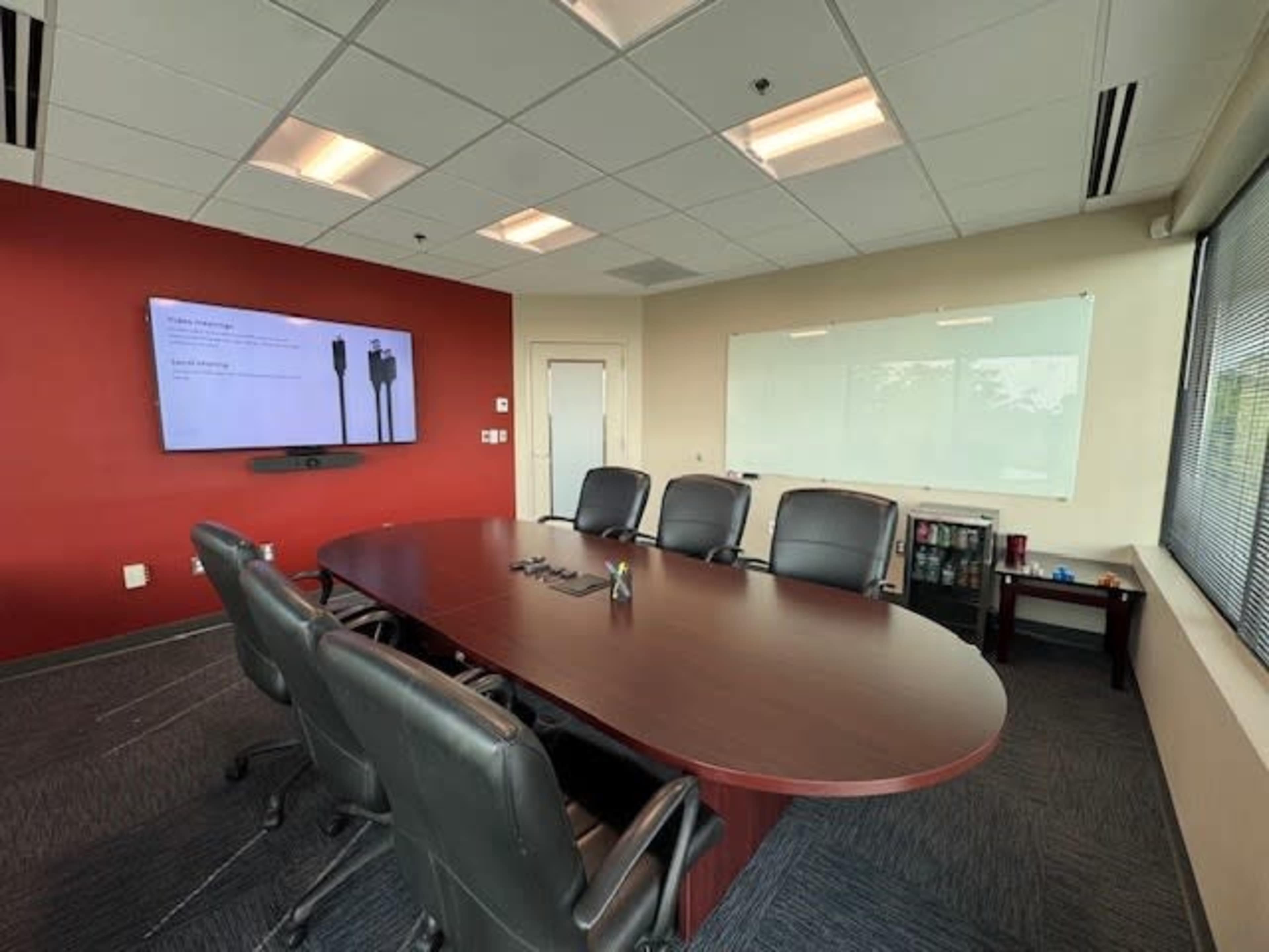 A modern conference room features a large wooden table surrounded by black chairs, with a screen displaying a presentation on one wall and a whiteboard on another.