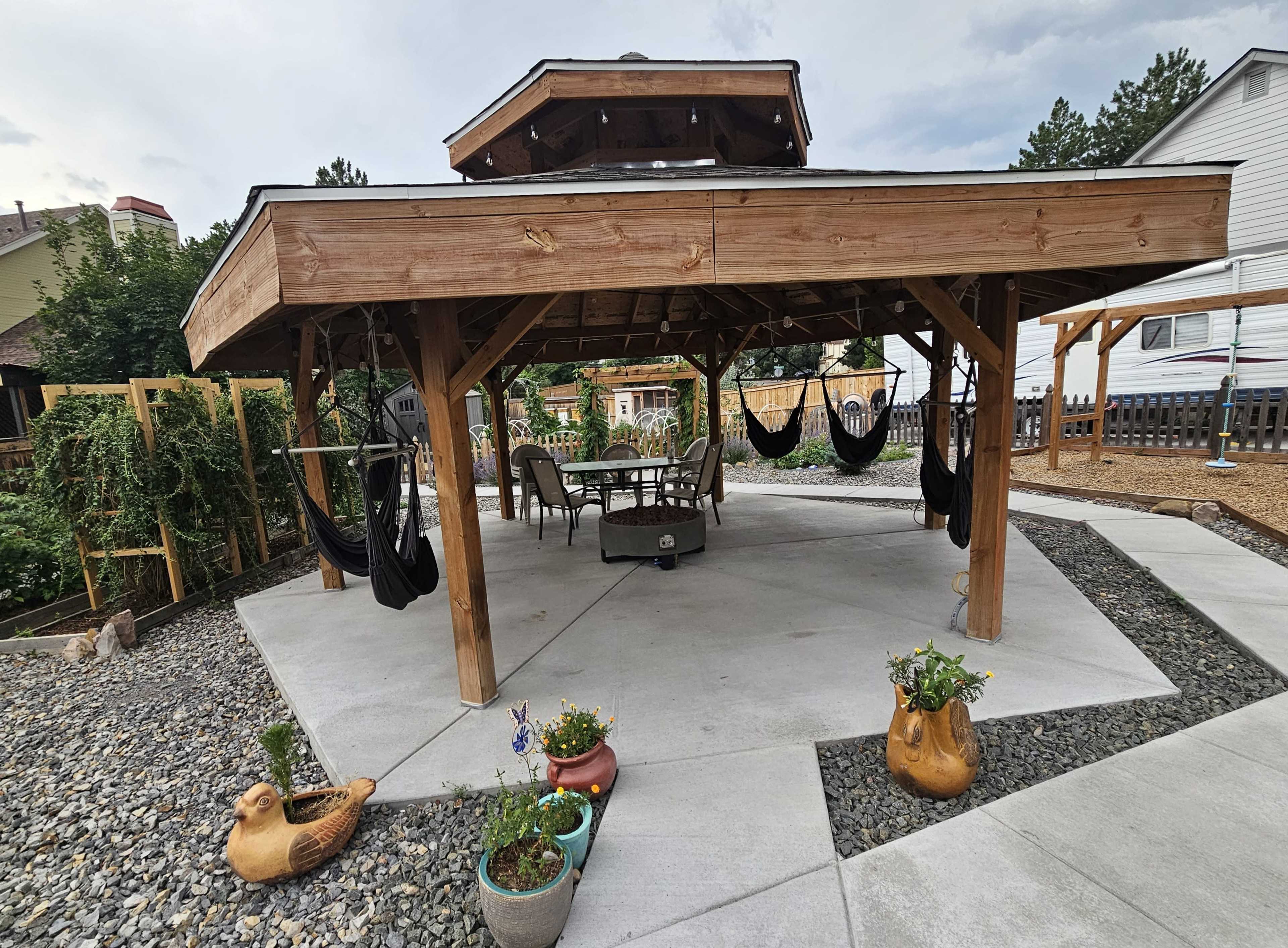 The image shows a wooden gazebo with a round roof, featuring a table and chairs underneath, surrounded by gravel and potted plants.