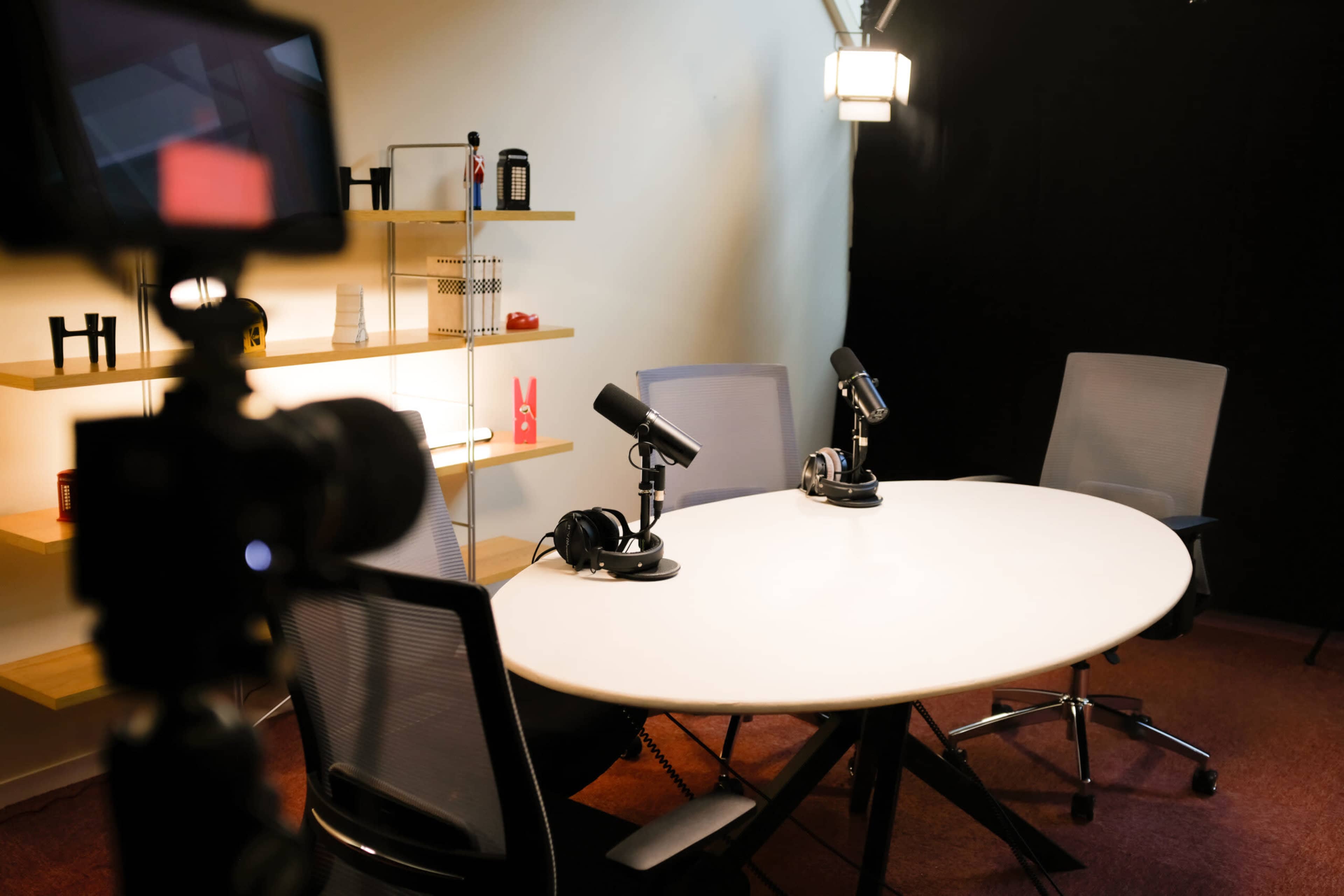 The image shows a podcast recording setup with a round table, two microphones, headphones, and shelves displaying various items in a brightly lit room.