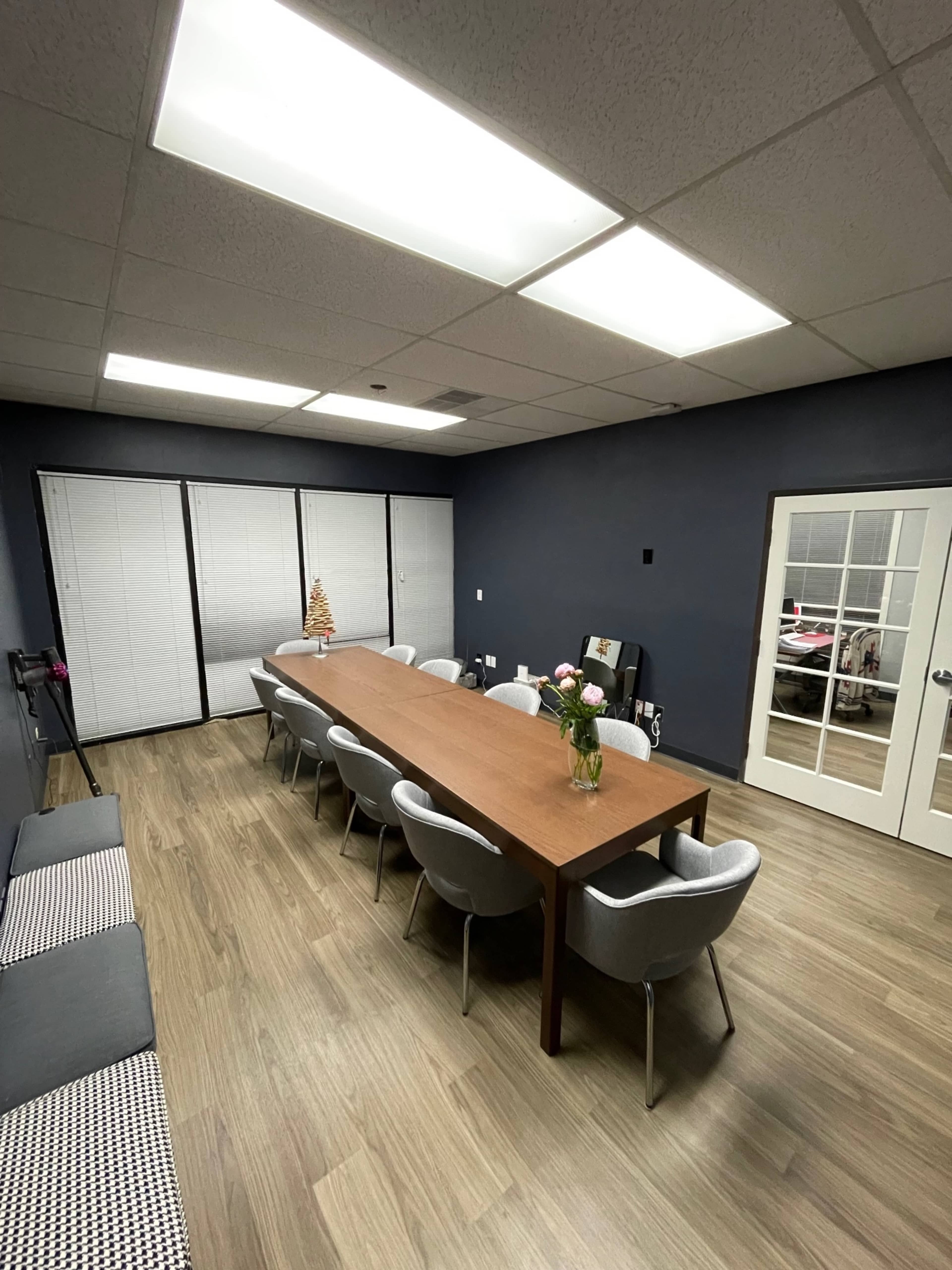 A long wooden table is surrounded by chairs in a well-lit conference room with dark blue walls and large windows covered by blinds.