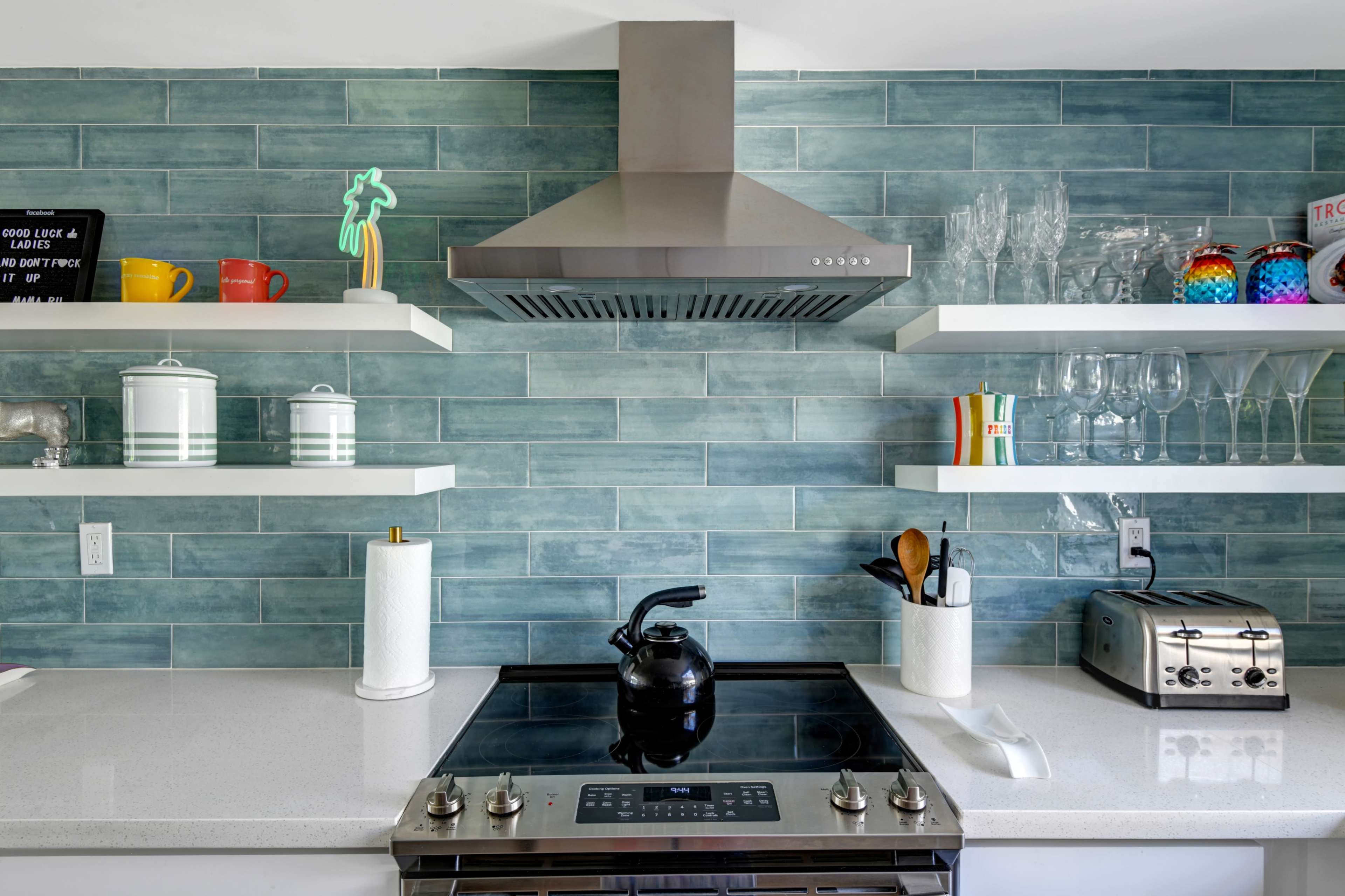 A modern kitchen features a stainless steel range hood above a cooktop, with teal tiled walls and open shelves displaying various kitchen items.