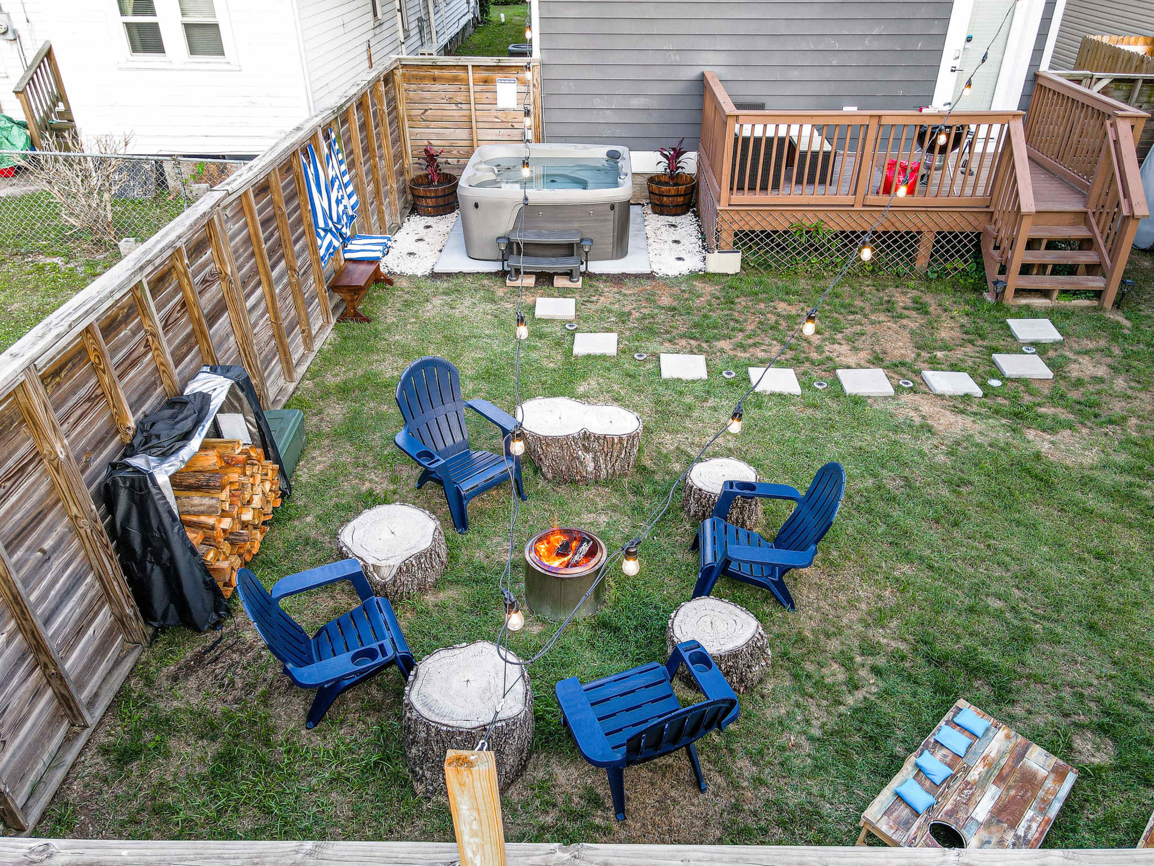 A backyard scene features circular seating made from tree stumps, a fire pit in the center, and a hot tub surrounded by decorative lights and wooden decking.
