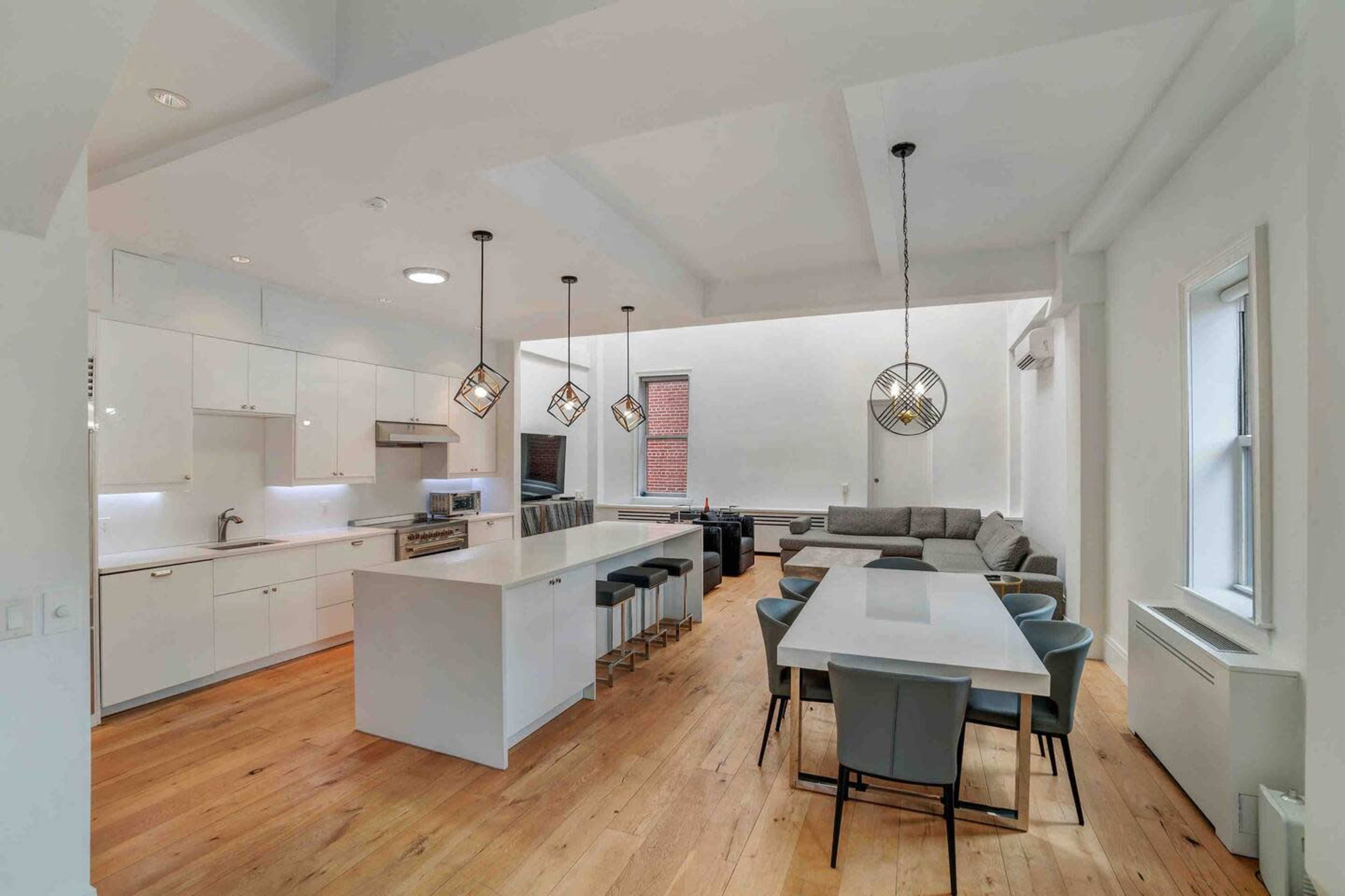 A modern kitchen and dining area with a large island, sleek cabinetry, a dining table, and a sectional sofa in a well-lit space.