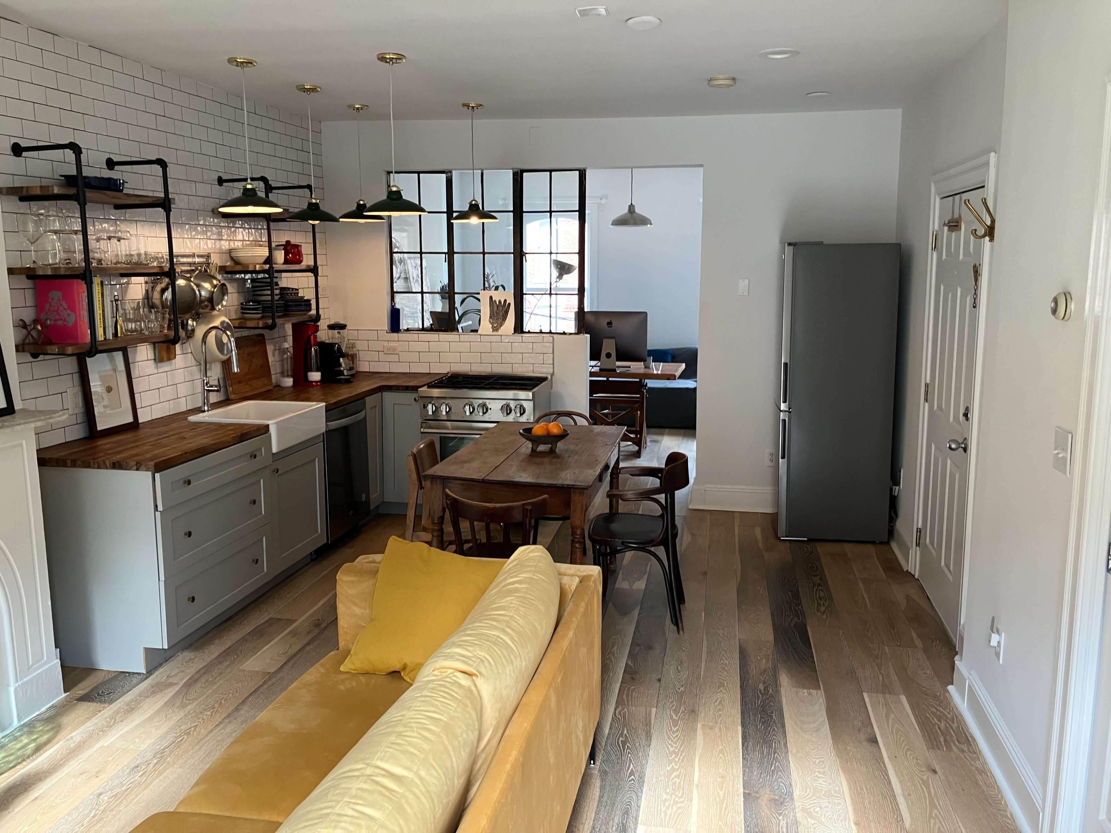 The image shows a modern kitchen and living area with wooden floors, a dining table, a refrigerator, and industrial-style lighting fixtures.