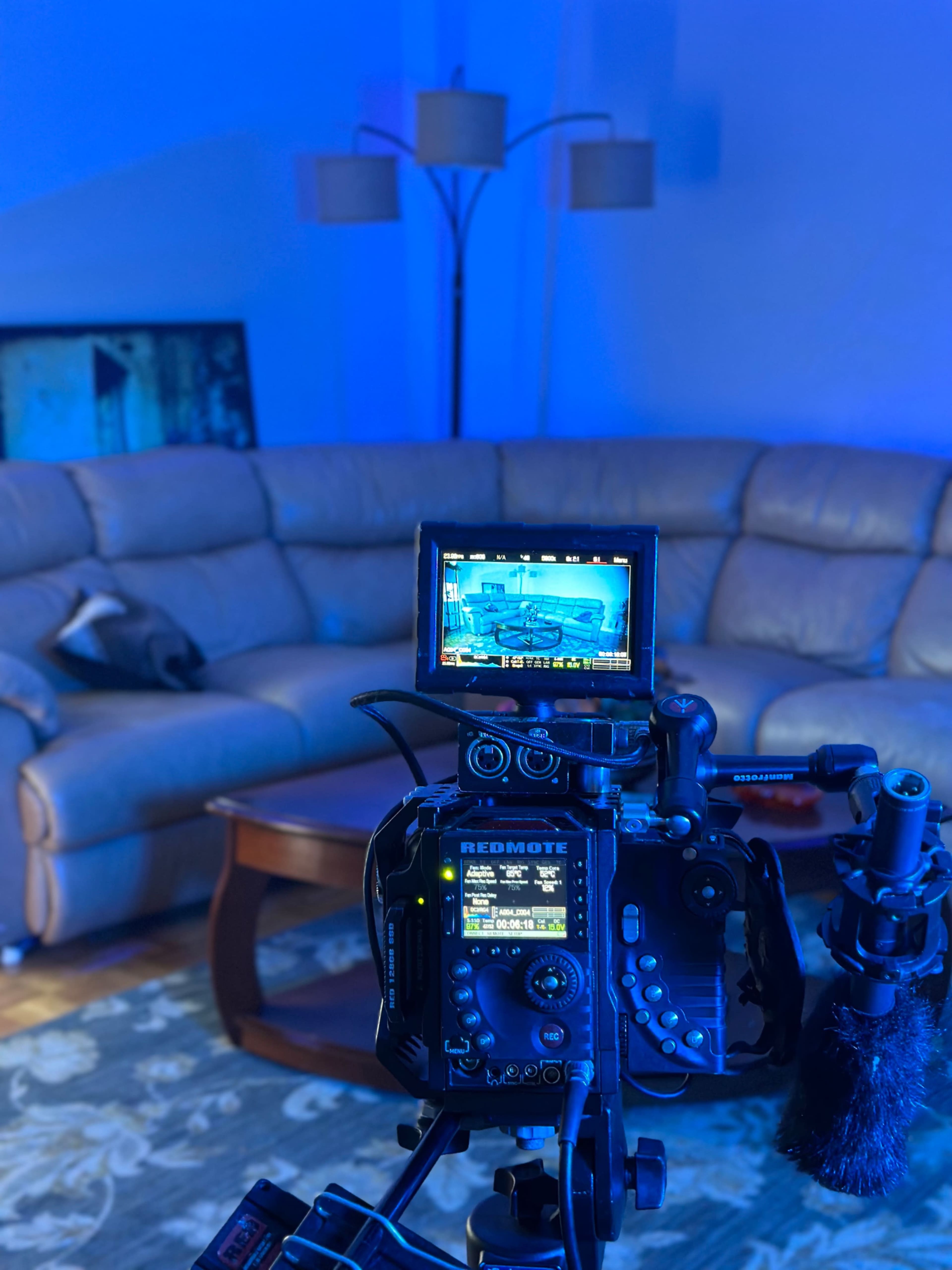 A camera on a tripod points at a well-lit living room scene featuring a sofa, a coffee table, and a floor lamp.