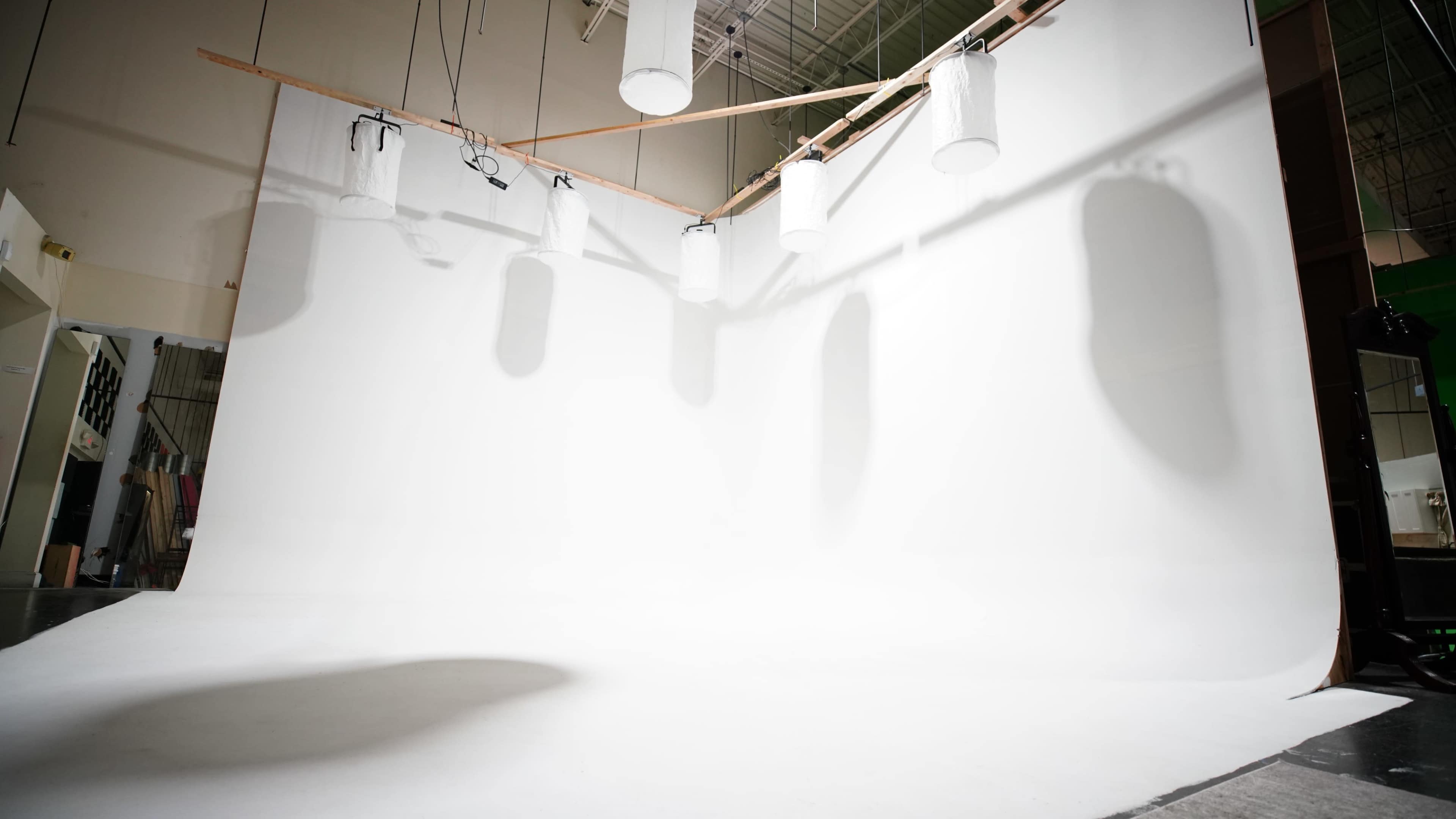 A spacious photography studio features a white backdrop with several hanging lights and shadows on the floor.