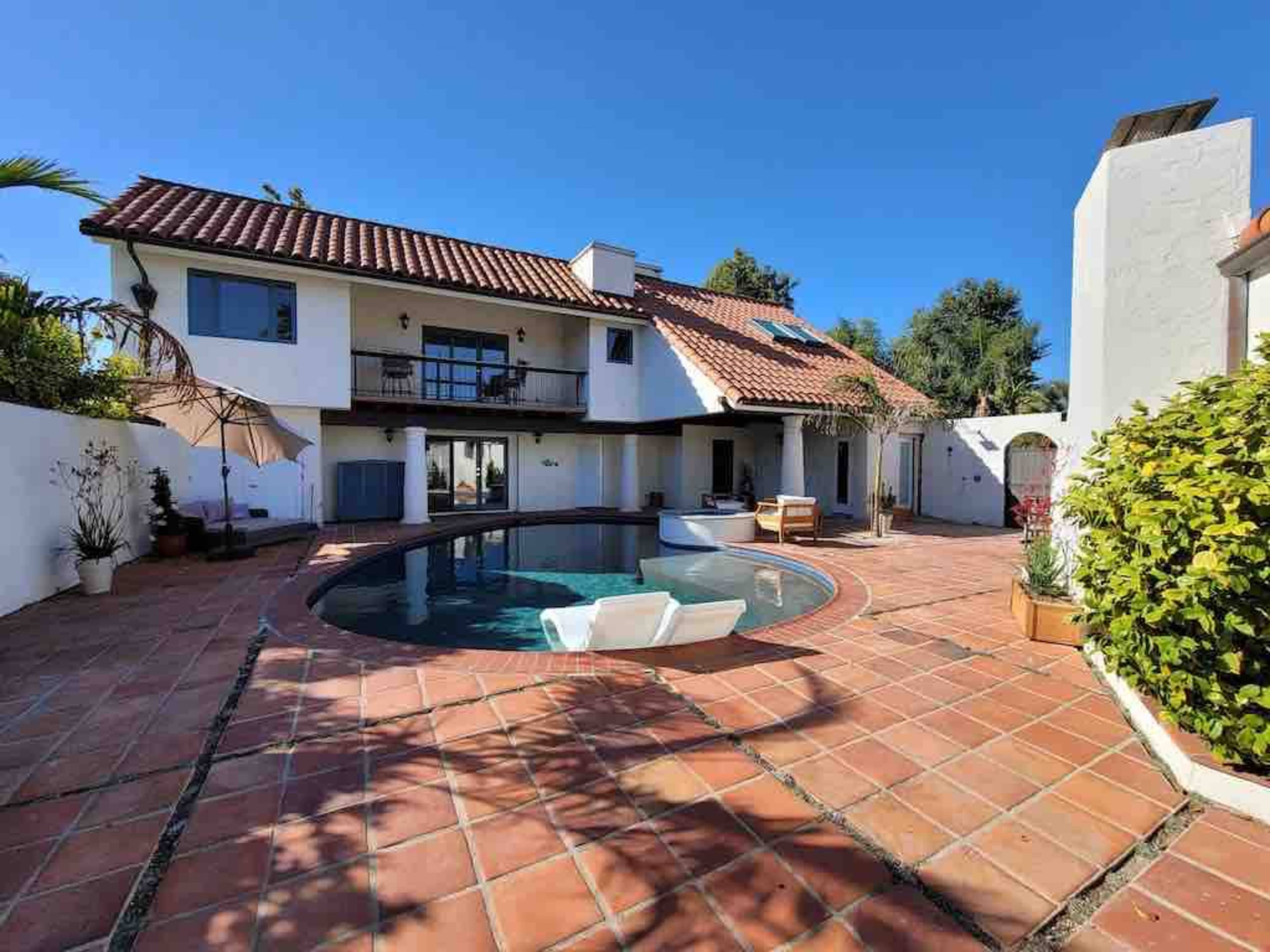 A house with a red-tiled roof surrounds a circular swimming pool, surrounded by a paved patio and landscaped greenery.