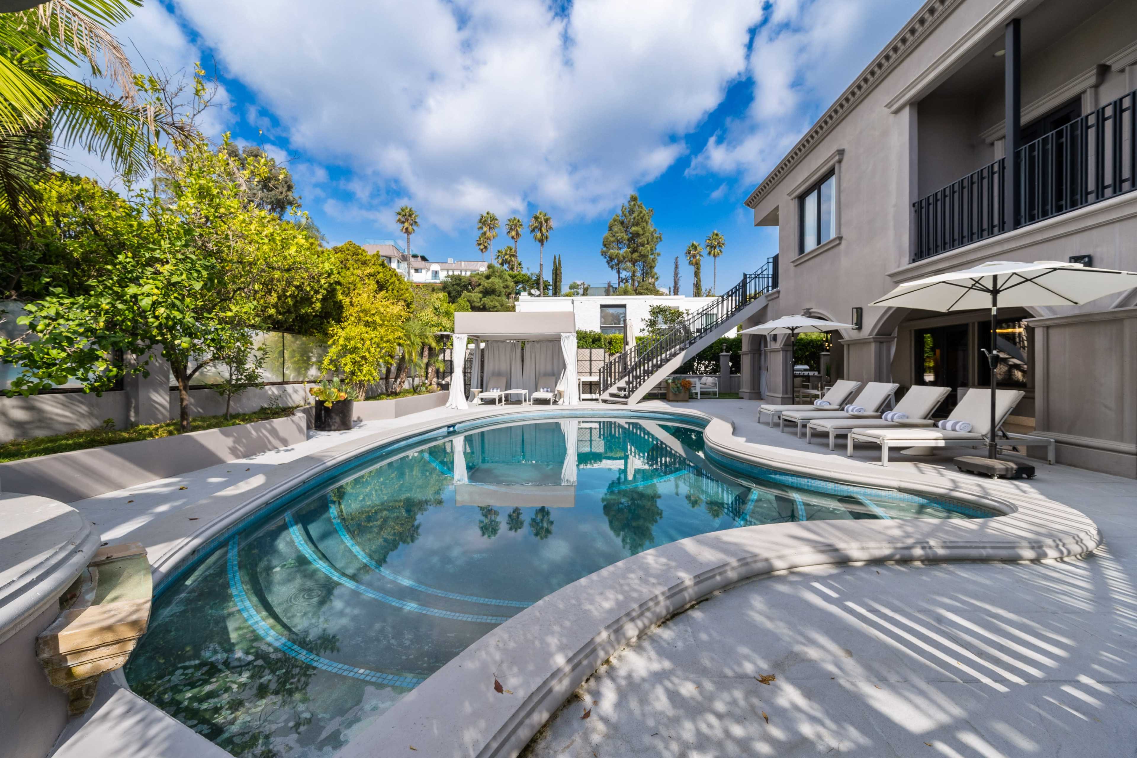 The image depicts a modern backyard with a curved swimming pool surrounded by lounge chairs and lush greenery.