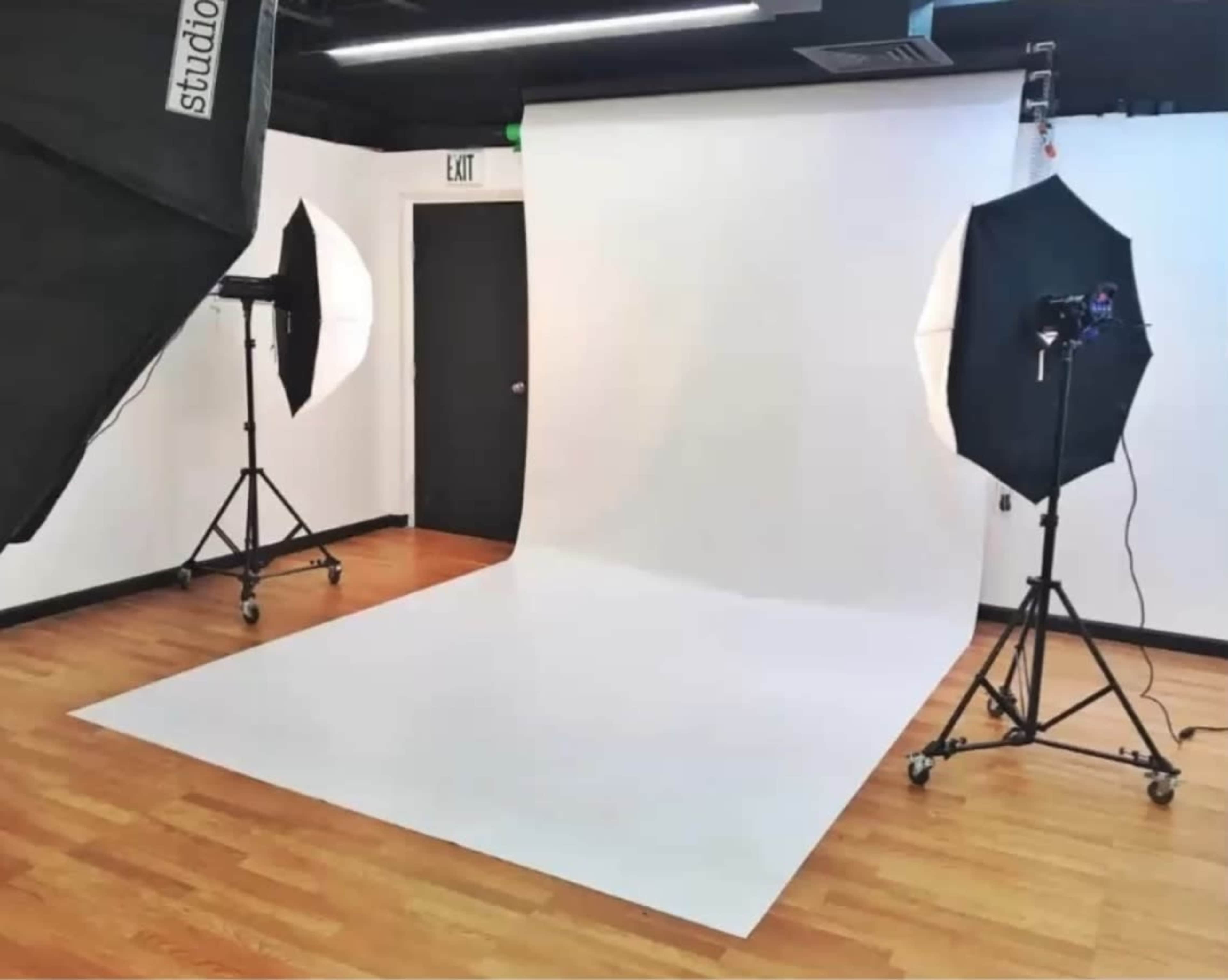 The image shows a photography studio with a white backdrop, two softbox lights, and studio equipment positioned on a wooden floor.