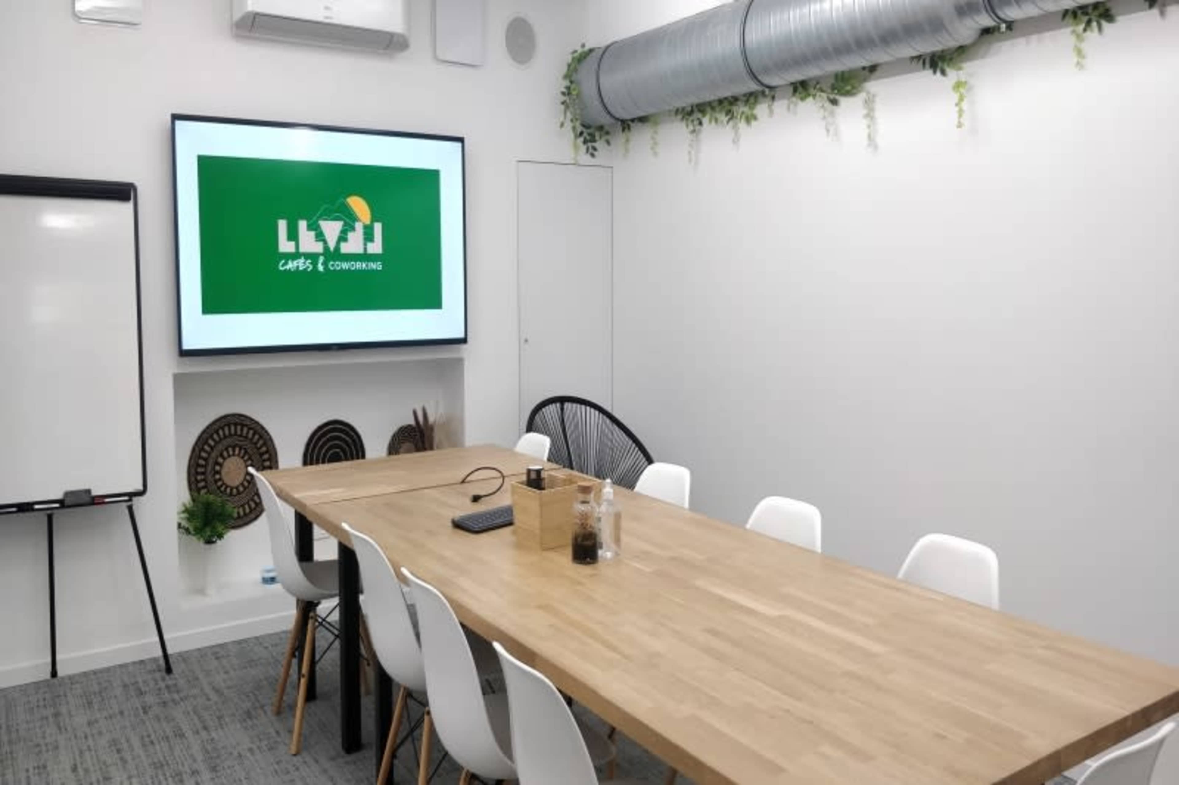 The image shows a modern conference room featuring a large wooden table surrounded by white chairs, with a screen displaying a green logo on the wall.