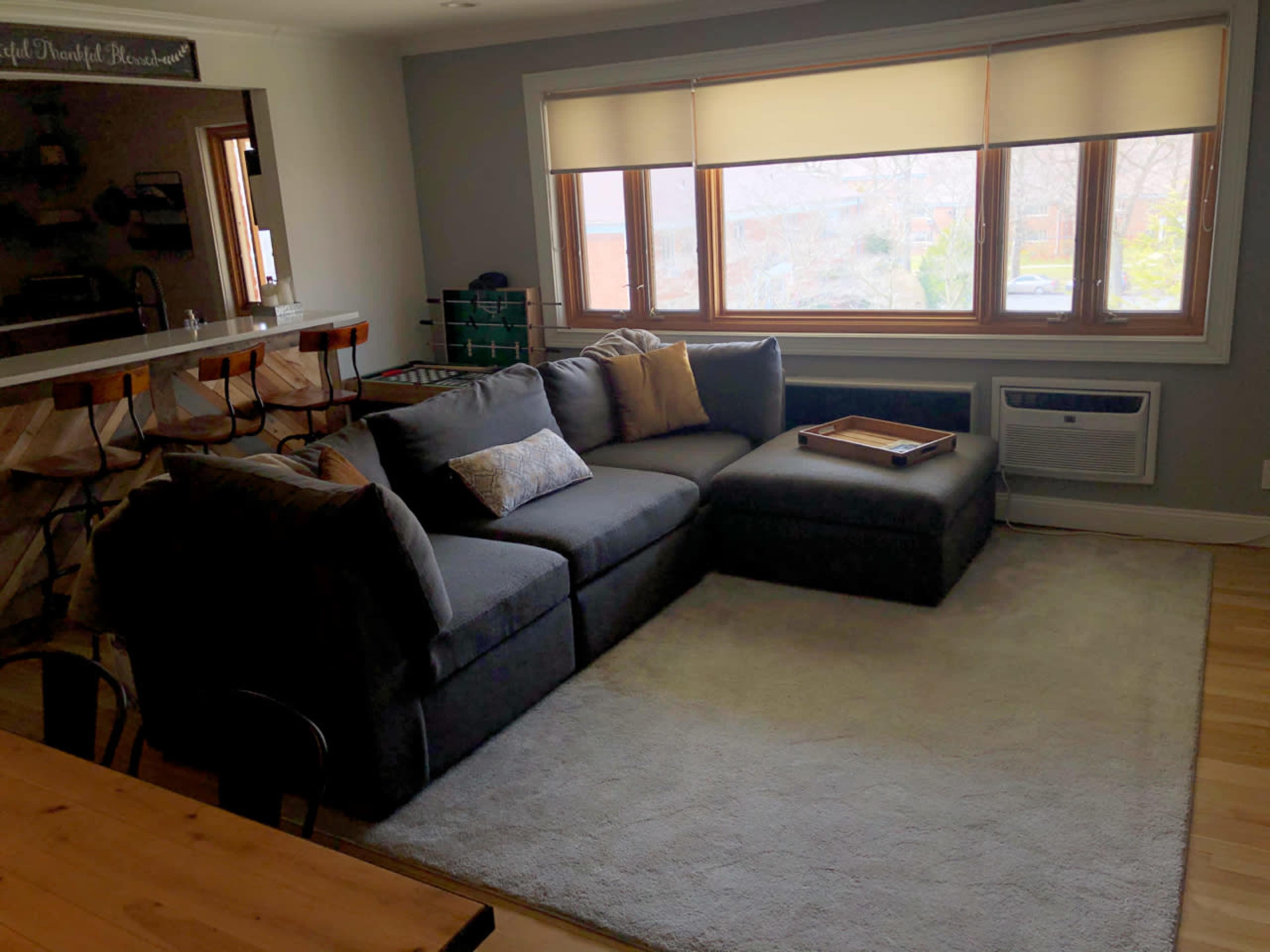 The image shows a modern living room with a large sectional sofa, a coffee table on a rug, and a window revealing an outdoor view.